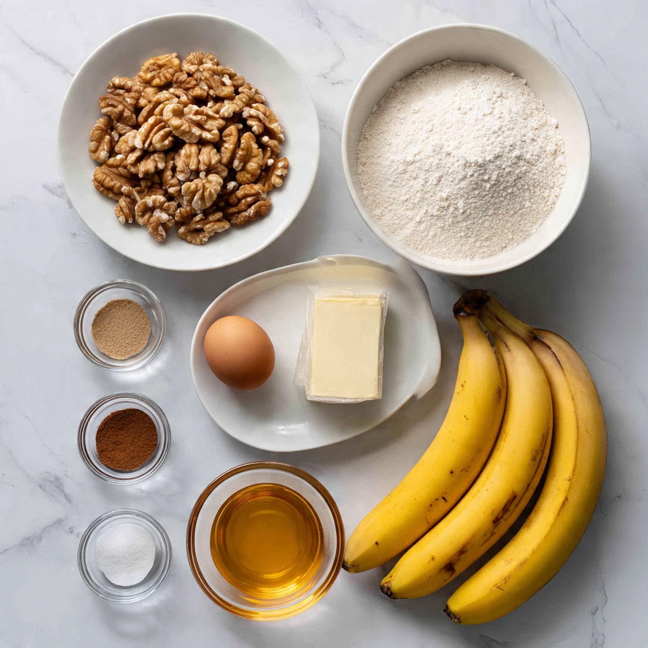 The image shows ingredients arranged on a white marbled surface. There are three ripe yellow bananas with brown spots placed horizontally at the bottom right. Above the bananas, there is a small wrapped block of butter and a single brown egg. To the left, there is a white bowl filled with light brown walnut halves. On the top right, a larger white bowl is filled with white flour, and next to it on the right, a smaller white plate holds white granulated sugar. Below these, there are four small glass bowls arranged in a square: the top left contains brown ground cinnamon, the top right holds a small amount of white salt, the bottom left has baking soda, and the bottom right contains baking powder. Below these glass bowls is another small glass bowl filled with a golden liquid, likely honey or syrup. photo taken with an iphone --ar 4:5 --v 7