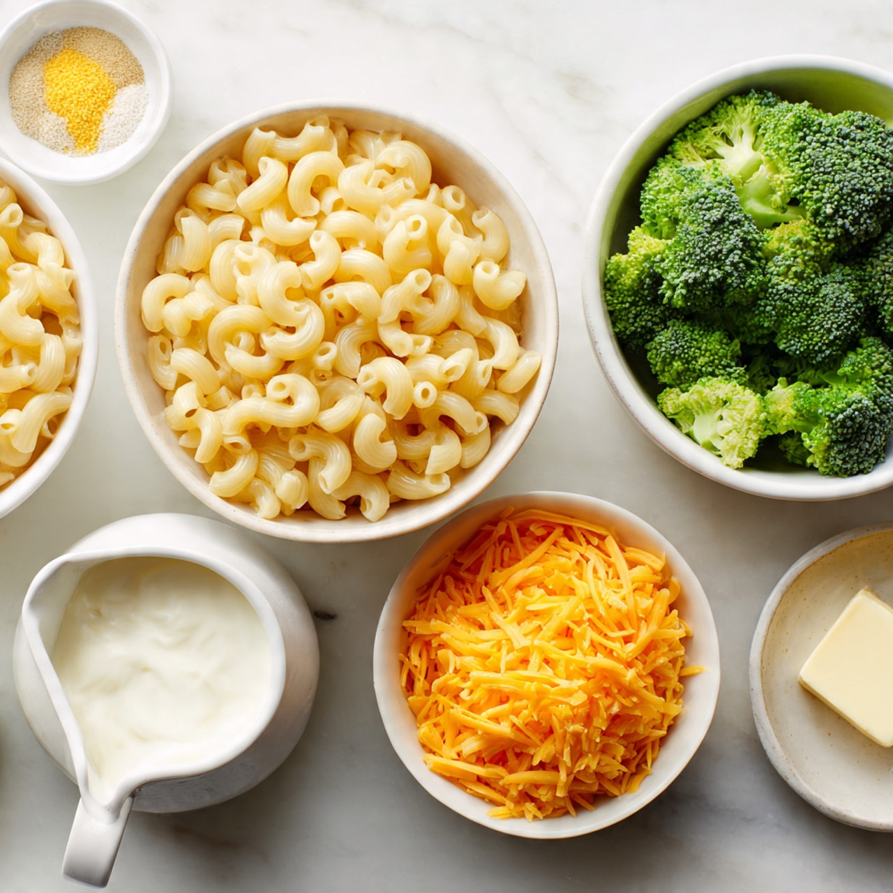 Five small white bowls are placed on a white marbled surface. The largest bowl holds short elbow macaroni pasta, pale yellow and slightly shiny, filling the bowl fully. Next to it, a bowl contains fresh green broccoli florets with a textured surface. Another bowl is filled with bright orange shredded cheese, soft and slightly fluffy in appearance. A smaller bowl has a soft white creamy cheese or cream, smooth and thick. On the top left, a small bowl holds yellow seasoning powder and white granulated salt mixed together. There is also a small white jug with a creamy white liquid, and a tiny white dish holds a small square of pale butter. The scene is bright and evenly lit, photo taken with an iphone --ar 4:5 --v 7