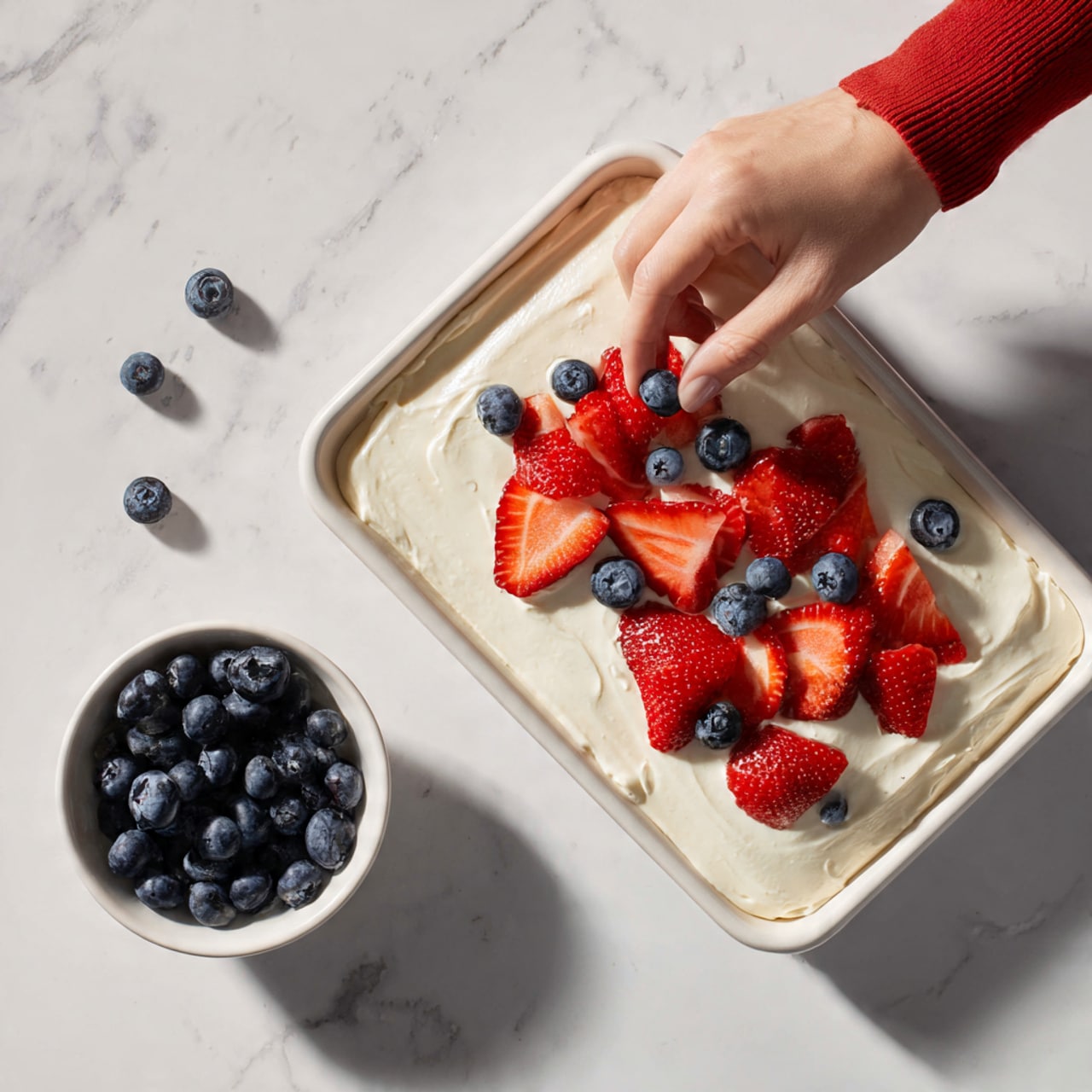A rectangular white tray holds a dessert with a thick, smooth white cream layer spread evenly on top. Scattered over the cream are bright red strawberry slices and round dark blue blueberries, spread across the surface in a loose pattern. A woman's hand wearing a red sleeve is seen placing a blueberry on the cream. Nearby, a small white bowl with additional blueberries rests on a white marbled surface. The scene is light and clean, emphasizing the fresh, vibrant fruit on the creamy layer photo taken with an iphone --ar 4:5 --v 7