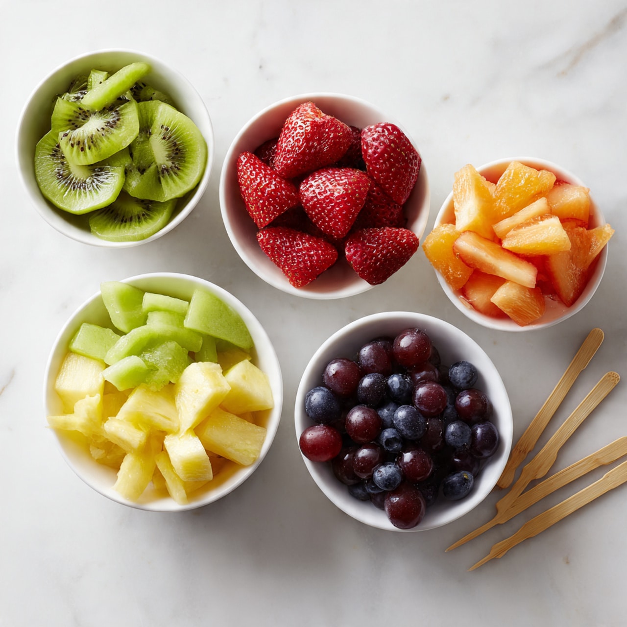 Five small white bowls are arranged on a white marbled surface in a loose cluster. Each bowl contains a different colorful fruit: one with bright green sliced kiwi, one with whole deep red strawberries, one with pale yellow pineapple chunks, one with orange cantaloupe pieces, and the last with a mix of purple grapes and dark blueberries. Wooden picks are placed beside some of the bowls. The image is bright and fresh, with the clean white bowls and marbled surface making the colorful fruits stand out. photo taken with an iphone --ar 4:5 --v 7