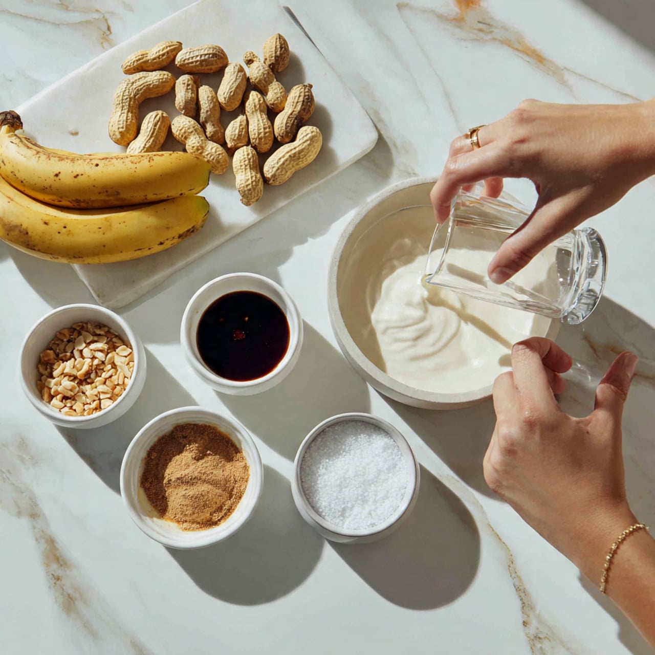 The image shows a white marbled surface with clusters of peanut shells and three bananas at the top left. Below the bananas, there are small white bowls arranged with different ingredients: one bowl has a dark brown powder, another with a light brown powder, a third bowl holds a dark syrup, and a larger bowl contains white granulated sugar. To the right, a woman's hand is holding a clear glass pitcher, pouring white liquid, likely milk, into the larger bowl of sugar. The scene is bright and clean, with soft natural light. photo taken with an iphone --ar 4:5 --v 7