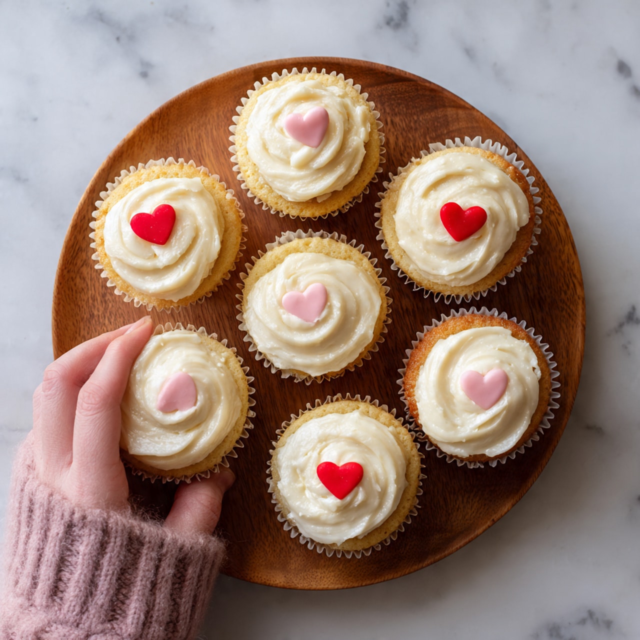 A round wooden plate holds eight small, smooth white frosted cupcakes. Each cupcake has two small heart-shaped decorations placed near the center, one red and one pink, on top of the glossy white icing. The cupcakes are arranged in a circle with one in the middle, all against a white marbled surface. A woman's hand reaches into the frame, touching one of the cupcakes lightly. Photo taken with an iphone --ar 4:5 --v 7
