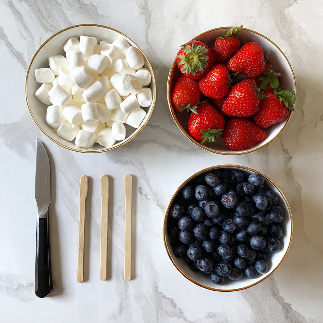 The image shows three white bowls with gold rims placed on a white marbled surface. The left bowl is filled with small white marshmallows, the middle bowl holds bright red strawberries with green stems, and the right bowl contains dark blue blueberries. Below the bowls, there is a black knife with a silver blade and five wooden skewers arranged in a row. Photo taken with an iphone --ar 4:5 --v 7