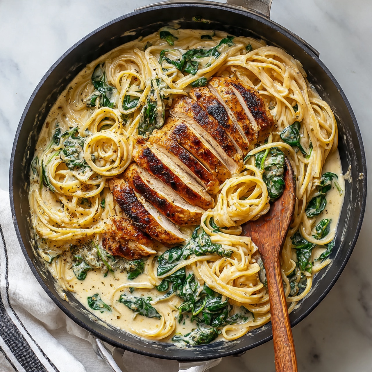 A black skillet sits on a white marbled surface holding a creamy pasta dish with three sliced pieces of brown, grilled chicken arranged in a semicircle on the top right. The pasta noodles are long and light yellow, mixed with wilted, dark green spinach leaves soaked in a creamy light beige sauce. A wooden spoon rests inside the pan on the right side, touching the food. The pan handle extends to the upper right corner. A white cloth with black stripes is partly visible at the bottom left corner. Photo taken with an iphone --ar 4:5 --v 7