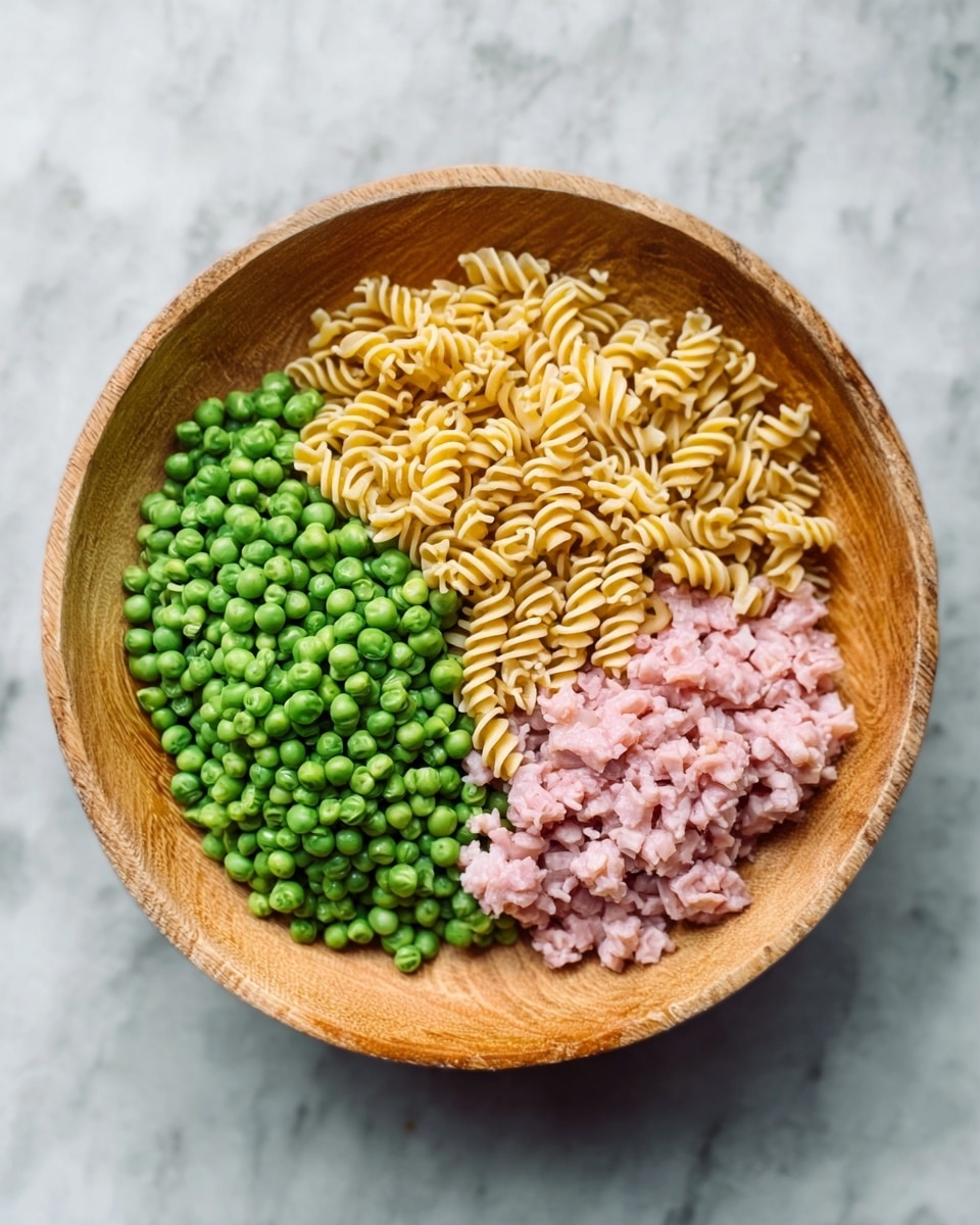 A wooden bowl contains three sections of food arranged in neat, separate layers. On the left is a pile of bright green peas with a fresh, round texture. On the right is a group of finely chopped pale pink pieces, likely some meat. The top section holds twisted, dry pasta pieces that are light yellow. The bowl sits on a white marbled surface. The photo was taken with an iphone --ar 4:5 --v 7