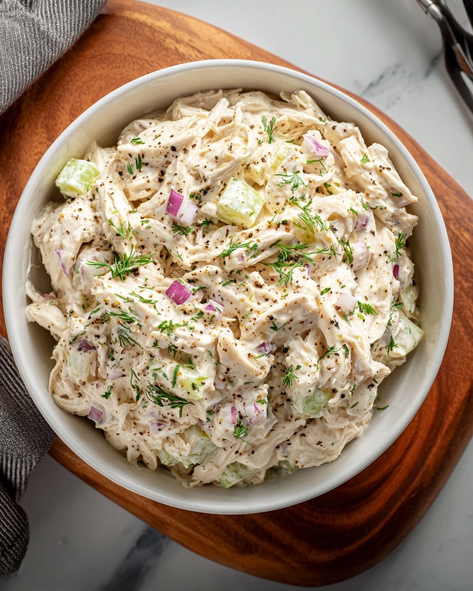 The dish is a creamy salad served in a round white bowl, filled with about three main layers: shredded chicken coated with a thick white creamy sauce, small chunks of pale green celery mixed evenly, and tiny pieces of red onion scattered throughout adding a slight purple color. The surface is sprinkled with black pepper and small green herbs, giving texture and color contrast. The bowl sits on a wooden board with a white marbled surface partially visible underneath. Photo taken with an iphone --ar 4:5 --v 7