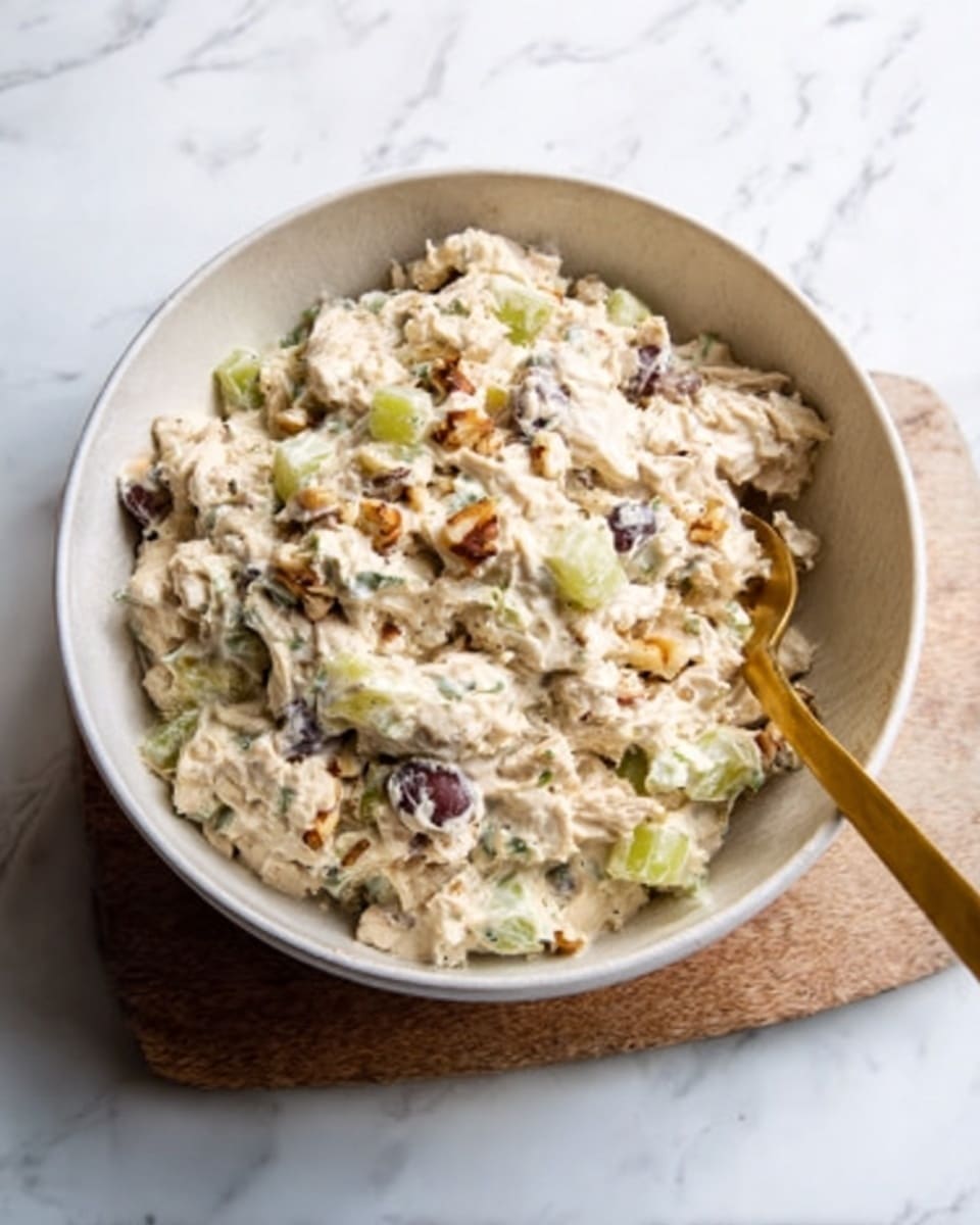 The image shows a white bowl filled with creamy chicken salad, with visible chunks of chicken mixed with small pieces of celery, nuts, and grapes. The salad has a light texture with a mix of beige, green, and purple colors spread evenly inside the bowl. A golden spoon is placed inside the bowl on the right side, partially submerged in the salad. The bowl is set on a wooden board placed on a white marbled surface, giving a clean and fresh look. Photo taken with an iphone --ar 4:5 --v 7
