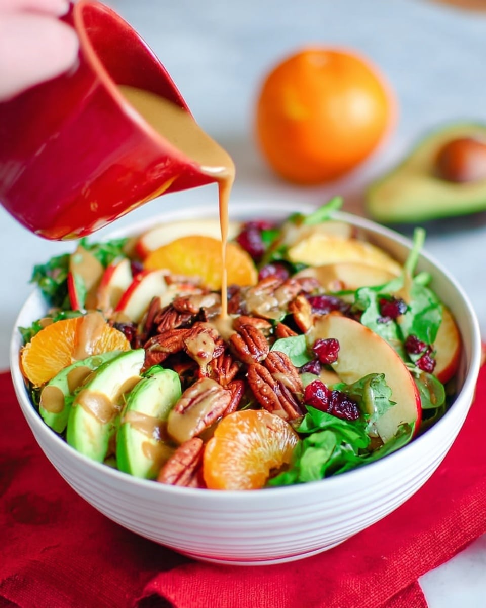 A white bowl filled with a colorful salad placed on a white marbled surface with a red cloth underneath, showing layers of orange mandarin slices, green avocado chunks, red apple slices, leafy spinach, dried cranberries, and pecans evenly mixed. A woman's hand is holding a small red cup pouring creamy brown dressing over the top, with a soft blurred background showing two whole mandarins and an avocado half. photo taken with an iphone --ar 4:5 --v 7