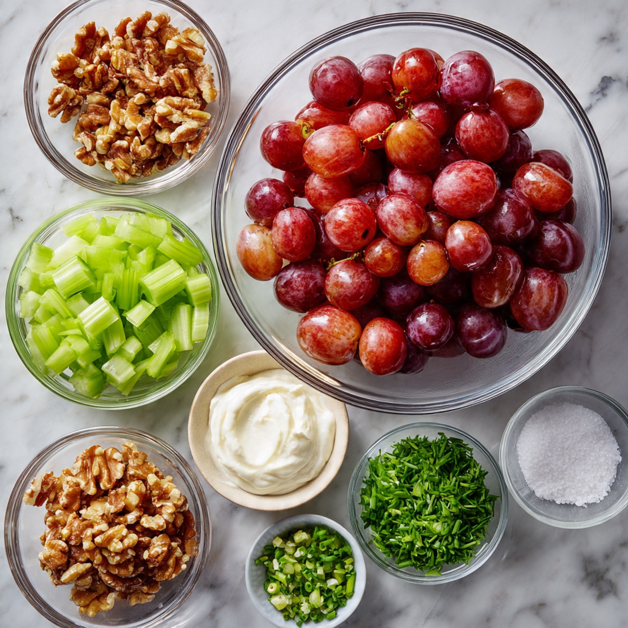 The image shows several clear glass bowls arranged on a white marbled surface, each containing different ingredients. One large bowl in the center is filled with halved red grapes, showing their glossy skin and juicy interiors. Surrounding it are smaller bowls holding bright green chopped celery, roughly chopped walnuts with their crunchy texture, finely chopped chives with a deep green color, fresh celery leaves, smooth white sour cream, creamy mayonnaise, and a small bowl of coarse salt. The bowls create a neat, colorful display of fresh and natural ingredients. photo taken with an iphone --ar 4:5 --v 7