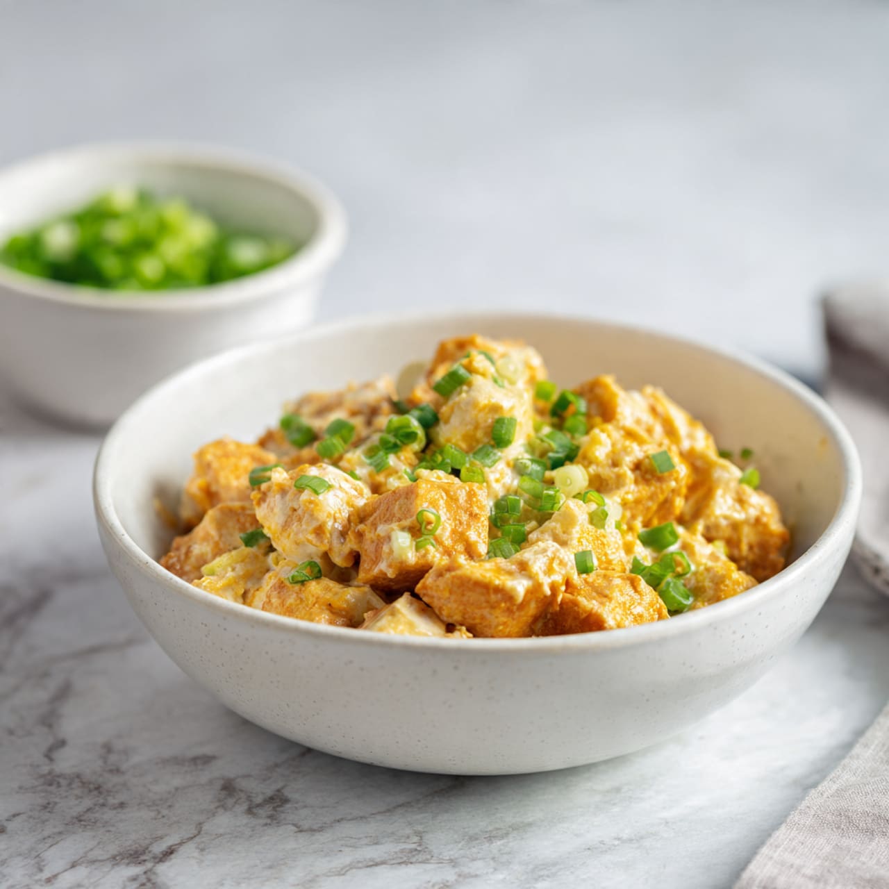 The image shows a food prep scene on a white marbled surface with an assortment of ingredients in white and light-colored bowls. There is a white tray on the left filled with small, cubed orange pieces of roasted squash. Above the tray, a white bowl contains fresh green spinach leaves. To the top center, a small white bowl holds light green pumpkin seeds, and a small glass with a bright green liquid is next to it. On the right side, a wooden board holds a white bowl filled with bright yellow diced mango, a white bowl with yellow corn kernels, black beans in a white bowl, and a small white bowl with pink pickled onions. A green avocado, cut in half, rests on the wooden board near the bowls. The overall arrangement is neat and colorful, highlighting fresh and varied ingredients ready for assembling a meal. photo taken with an iphone --ar 4:5 --v 7