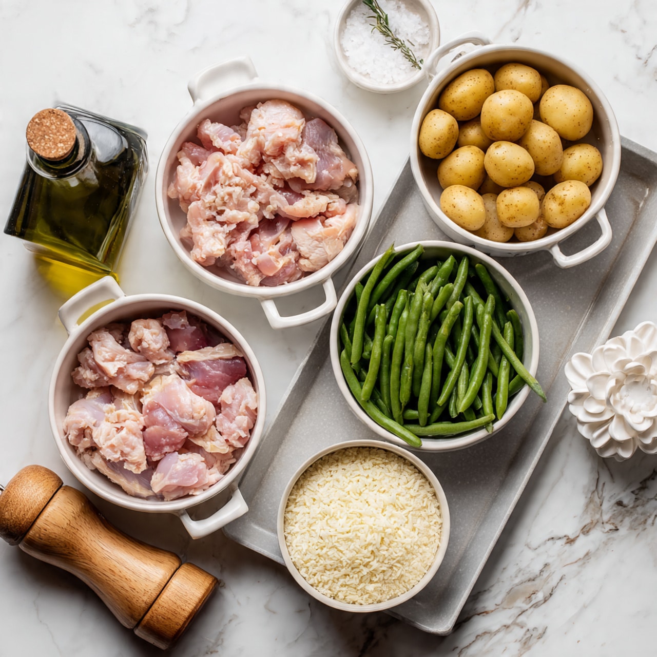 The image shows a baking tray with several white bowls placed on it, each containing different ingredients. One bowl has raw pink chicken pieces, another bowl filled with small round light brown potatoes, a third bowl holds green beans, and there is a smaller bowl with white rice or cheese grains. Next to the bowls, there is a dark green olive oil bottle, a wooden pepper grinder, and a small white bowl with salt. A white flower-shaped object is also visible near the chicken bowl. The whole setup is on a white marbled surface. Photo taken with an iphone --ar 4:5 --v 7