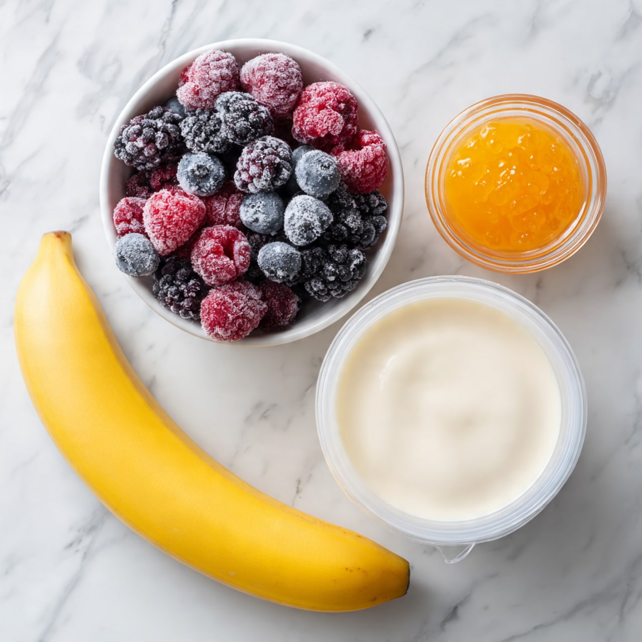 The image shows a white round bowl filled with mixed frozen berries including blackberries, raspberries, and blueberries, all covered lightly with frost. Next to it is a small transparent glass bowl holding an orange jelly or sauce. In front of these bowls is a white container filled with a smooth white substance, likely yogurt or cream. A yellow ripe banana with a slight curve lies at the bottom on a white marbled surface. A white measuring cup filled with milk is also present to the left. The overall scene is simple with fresh and creamy ingredients arranged neatly. Photo taken with an iphone --ar 4:5 --v 7