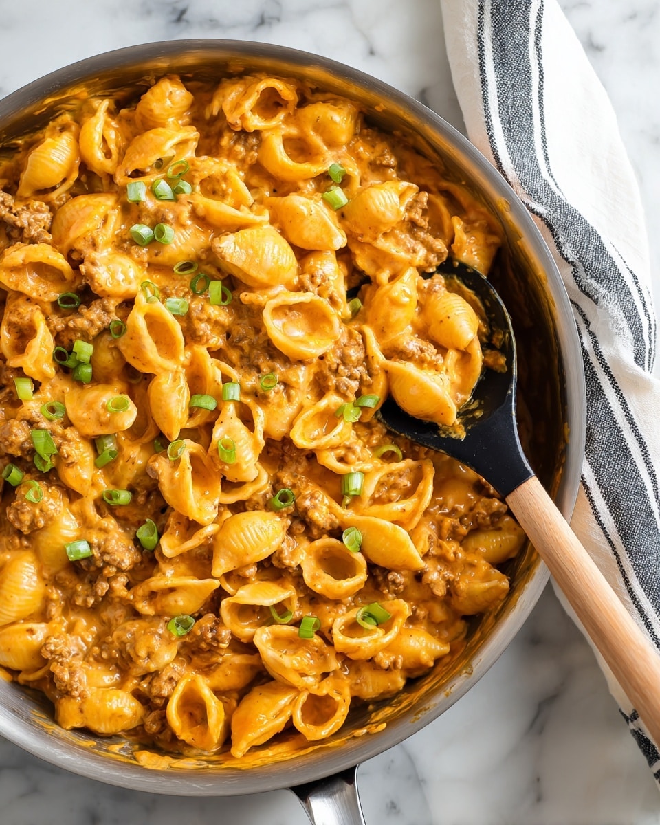 A close-up view of a creamy pasta dish in a silver pan sitting on a white marbled surface, showing one layer of small shell-shaped pasta coated in a thick orange cheese sauce mixed with small brown bits of ground meat and topped with scattered bright green chopped scallions. A black spoon with a light wooden handle scoops some pasta from the pan, lifting it slightly above the rest of the creamy pasta mixture. A white cloth with dark grey stripes is partially visible next to the pan photo taken with an iphone --ar 4:5 --v 7