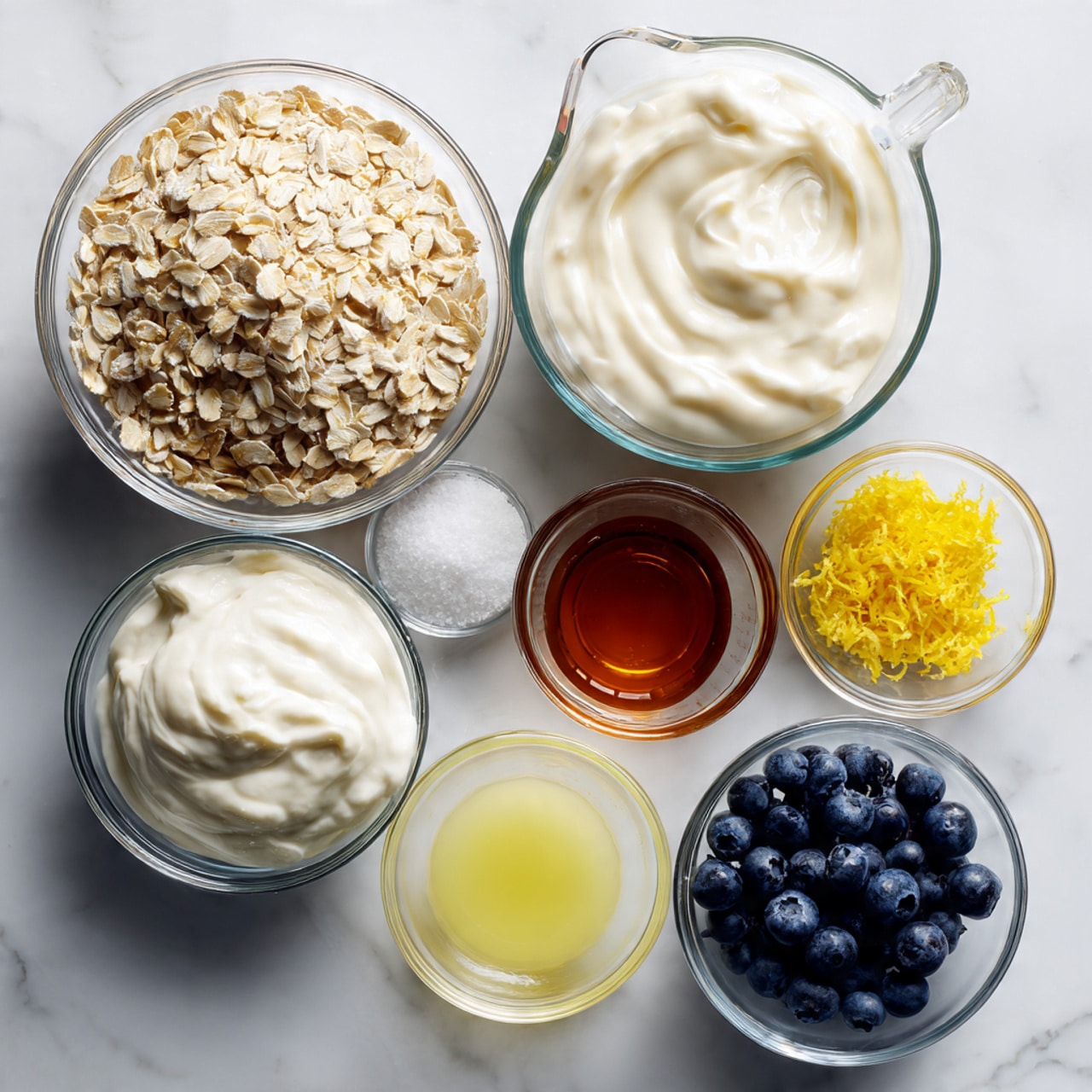 The image shows seven clear glass bowls and a clear glass measuring cup, all placed on a white marbled surface. The top left bowl is filled with light beige oats. Next to it on the right is a measuring cup with creamy white liquid. To the right of the measuring cup is a bowl filled with thick white yogurt. Below the oats is a small bowl with white salt. To the right and slightly down is a small bowl with pale yellow lemon juice. Next to it on the right is a small bowl with yellow lemon zest. In the center, slightly above, there is a small measuring cup with dark amber liquid, likely maple syrup, and to the right of it is a bowl filled with dark blue blueberries. The items are arranged in a loose circle, creating a balanced view of the ingredients. photo taken with an iphone --ar 4:5 --v 7