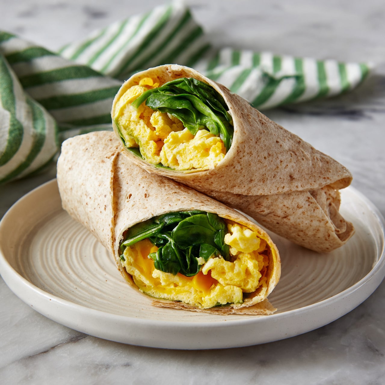 The image shows two halves of a wrap placed on a white round plate with subtle ridges. The wrap is light brown with a soft, textured surface. Inside, the wrap has three visible layers: bright yellow scrambled eggs with a fluffy texture, fresh green spinach leaves with a slightly crinkled look, and a hint of melted cheese that is light yellow and creamy. The plate rests on a white marbled surface with a folded striped green cloth nearby. photo taken with an iphone --ar 4:5 --v 7