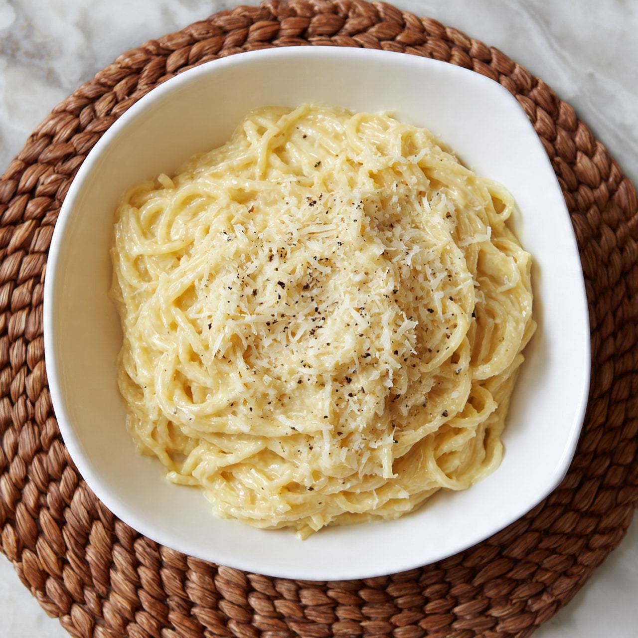 The image shows a white square bowl filled with creamy pasta, arranged in a circular mound in the center. The pasta is coated with a light yellow sauce that makes it look smooth and rich. On top, there is a thin layer of grated cheese creating a slightly rough white surface sprinkled with small, dark specks of black pepper. The bowl sits on a woven brown mat, all placed on a white marbled surface. Photo taken with an iphone --ar 4:5 --v 7