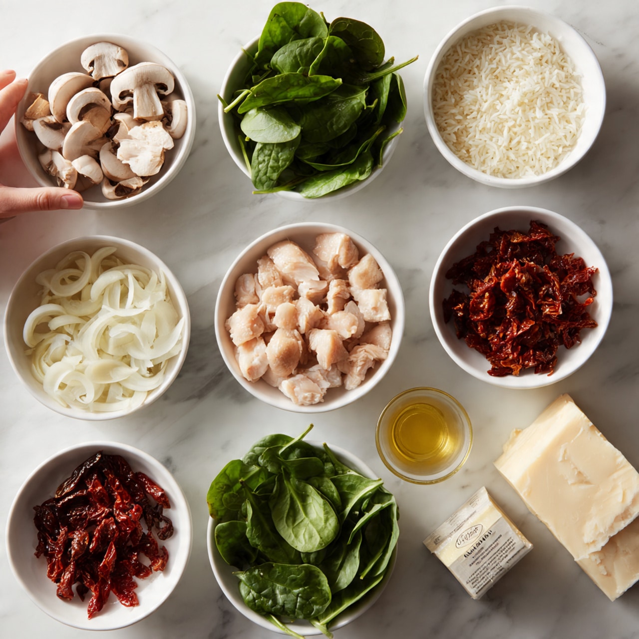 The image shows ingredients for cooking laid out on a white marbled surface. There are small white bowls filled with fresh green spinach leaves, sliced white mushrooms, chopped sun-dried tomatoes, chopped raw chicken pieces, sliced white onions, a light yellow liquid (likely oil), chopped garlic, cooked white rice, butter, and a small carton of Campbell's Real Stock chicken broth. Each bowl is neatly arranged with clear colors and textures visible in the ingredients. A woman's hand is slightly visible in the corner, about to pick up one of the bowls. The scene is bright and clean, ready for cooking photo taken with an iphone --ar 4:5 --v 7