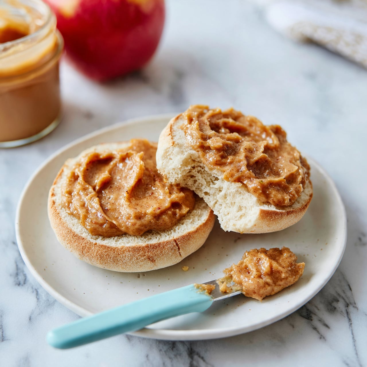 A white plate holds an open English muffin split into two halves; each half is topped with a thick layer of chunky, light brown apple cinnamon spread with visible small apple pieces. One half has a bite taken out of it, showing the soft, airy inside texture. A light blue spreader knife rests on the plate with a dollop of the same spread on its tip. The plate sits on a white marbled surface, with a red apple and a jar of spread partially visible in the background. photo taken with an iphone --ar 4:5 --v 7