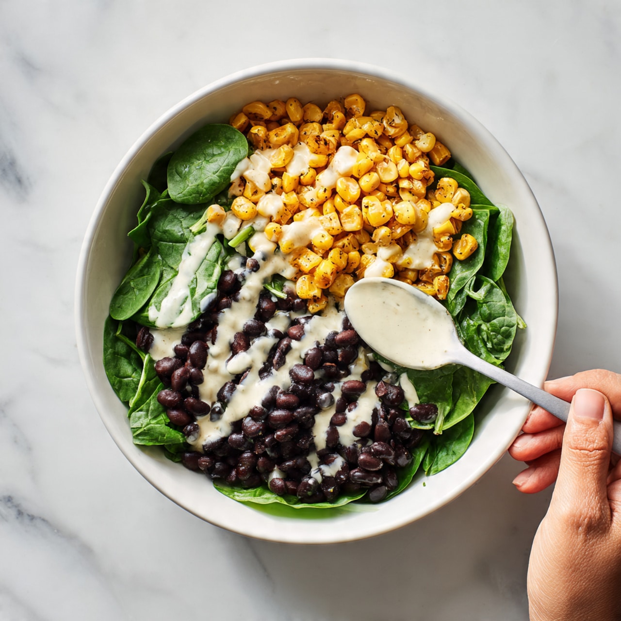 A white bowl with three main layers: at the bottom, fresh green spinach leaves spread evenly; on one side, a pile of small golden-brown roasted corn kernels with a slightly crispy texture; on the opposite side, a portion of shiny black beans; a creamy white sauce is drizzled over the middle, partly covering the spinach and beans; a woman's hand holds a white spoon resting inside the bowl; the bowl sits on a white marbled surface. photo taken with an iphone --ar 4:5 --v 7