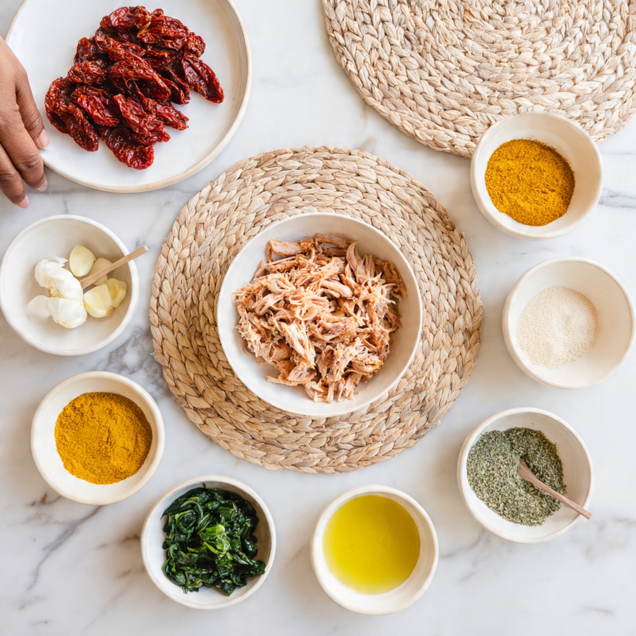 The image shows several small white bowls and dishes arranged neatly on a white marbled surface. A large white bowl filled with shredded light pink cooked chicken sits on a woven circular mat in the center. Around it are smaller white bowls containing various ingredients: bright green spinach leaves, pale yellow turmeric powder, a golden yellow liquid, white salt, light beige powder, and a small amount of garlic pieces. At the top left, a white plate holds deep red sun-dried tomatoes with a woman's hand reaching towards it. The arrangement is neat and colorful. photo taken with an iphone --ar 4:5 --v 7