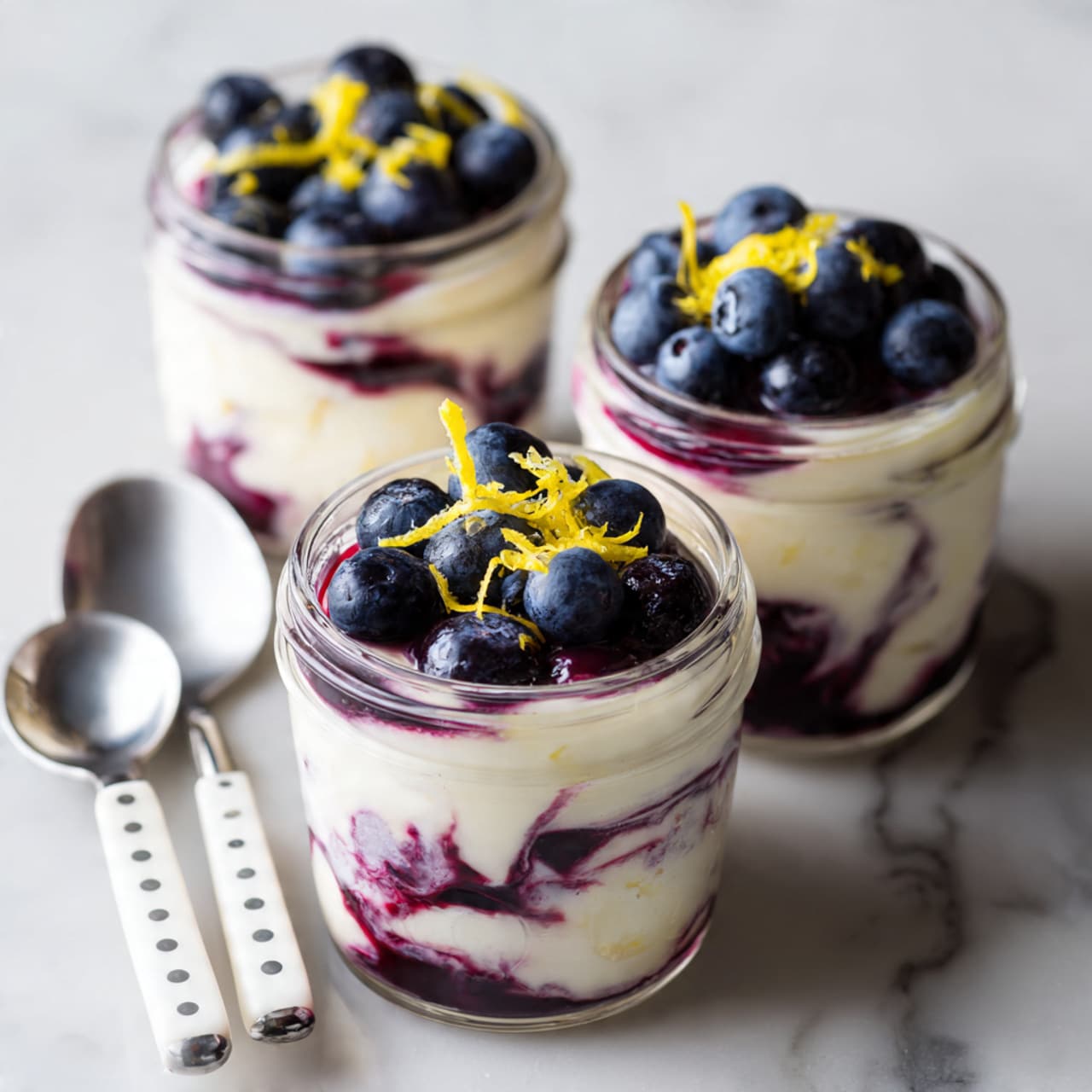 The image shows three small clear glass jars filled with layers of a creamy white base mixed with swirls of dark purple blueberry sauce. Each jar has a thick creamy white layer at the bottom, followed by a marbled mix of white and purple layers in the middle, topped with fresh dark blue blueberries and some yellow lemon zest on top. The jars are placed on a surface with a white marbled texture, and there are three silver spoons with white handles and gray dots lying next to the jars. photo taken with an iphone --ar 4:5 --v 7