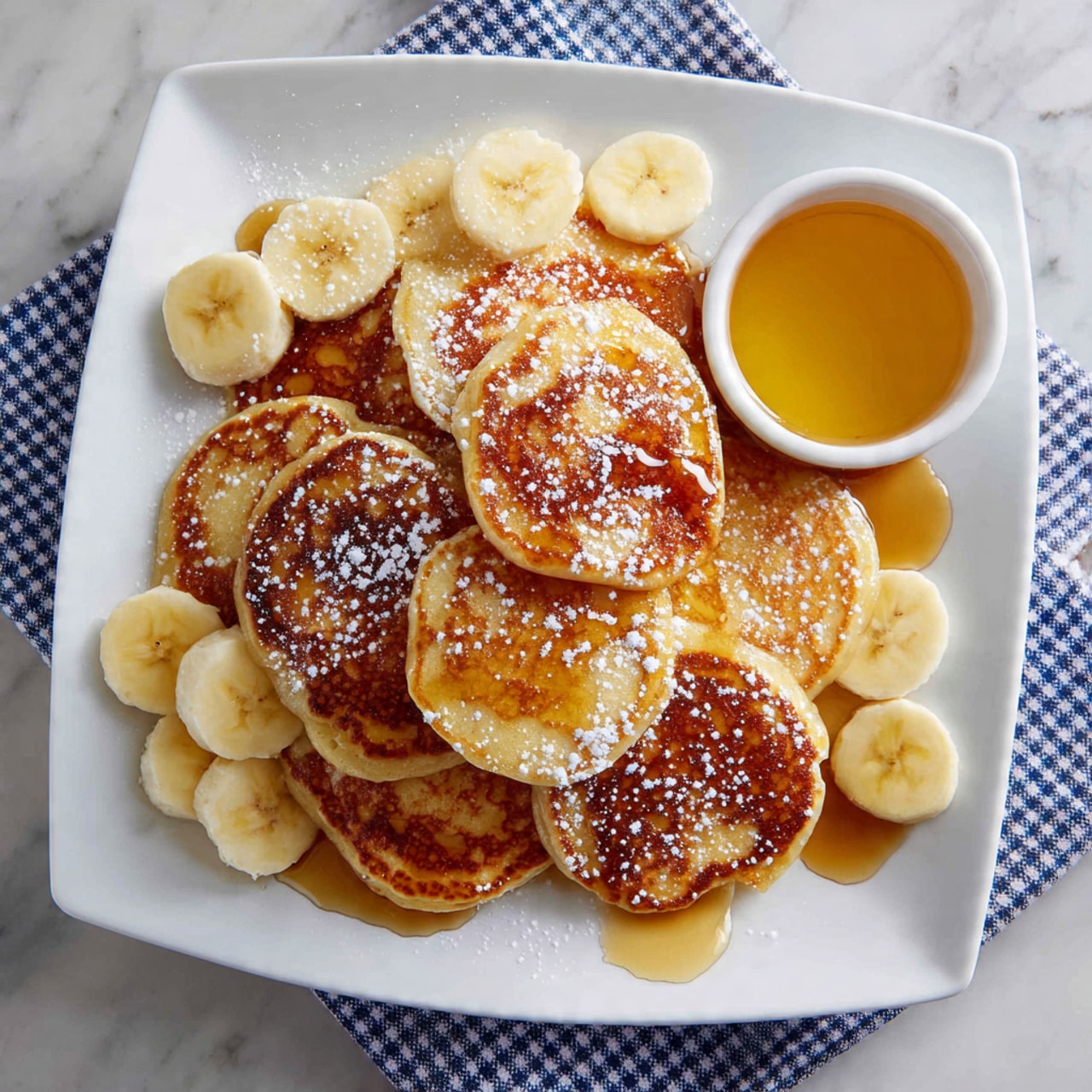 A white square plate filled with about 15 small golden-brown pancakes stacked loosely in the center, dusted with powdered sugar and drizzled with honey. On the plate’s sides are three peeled banana halves positioned around the pancakes. In the top right corner of the plate, there is a small white bowl filled with light golden syrup. The plate sits on a white marbled surface with a blue and white checkered cloth partially underneath. photo taken with an iphone --ar 4:5 --v 7
