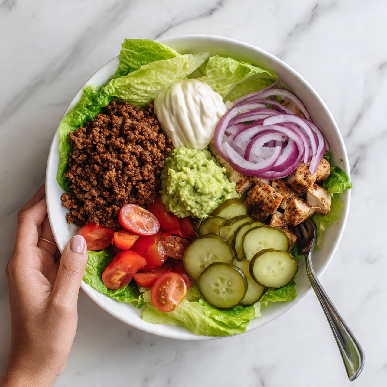 A white bowl sits on a white marbled surface filled with several colorful layers of food arranged clearly. At the base, fresh green lettuce leaves create a leafy texture. On one side, there is cooked ground meat with a crumbly texture in dark brown color. Next to it is a smooth dollop of pale cream-colored sauce, and beside that is a small portion of light green avocado mash. Grilled pieces of chicken with a browned, slightly charred look occupy another section. Thin rings of red onion are neatly stacked near the edge, alongside sliced round pickles which are light green with seeds visible inside. Bright red cherry tomatoes cut in halves complete the bowl, showing their juicy, smooth interiors. A silver fork rests inside the bowl with a woman's hand holding it. Photo taken with an iphone --ar 4:5 --v 7