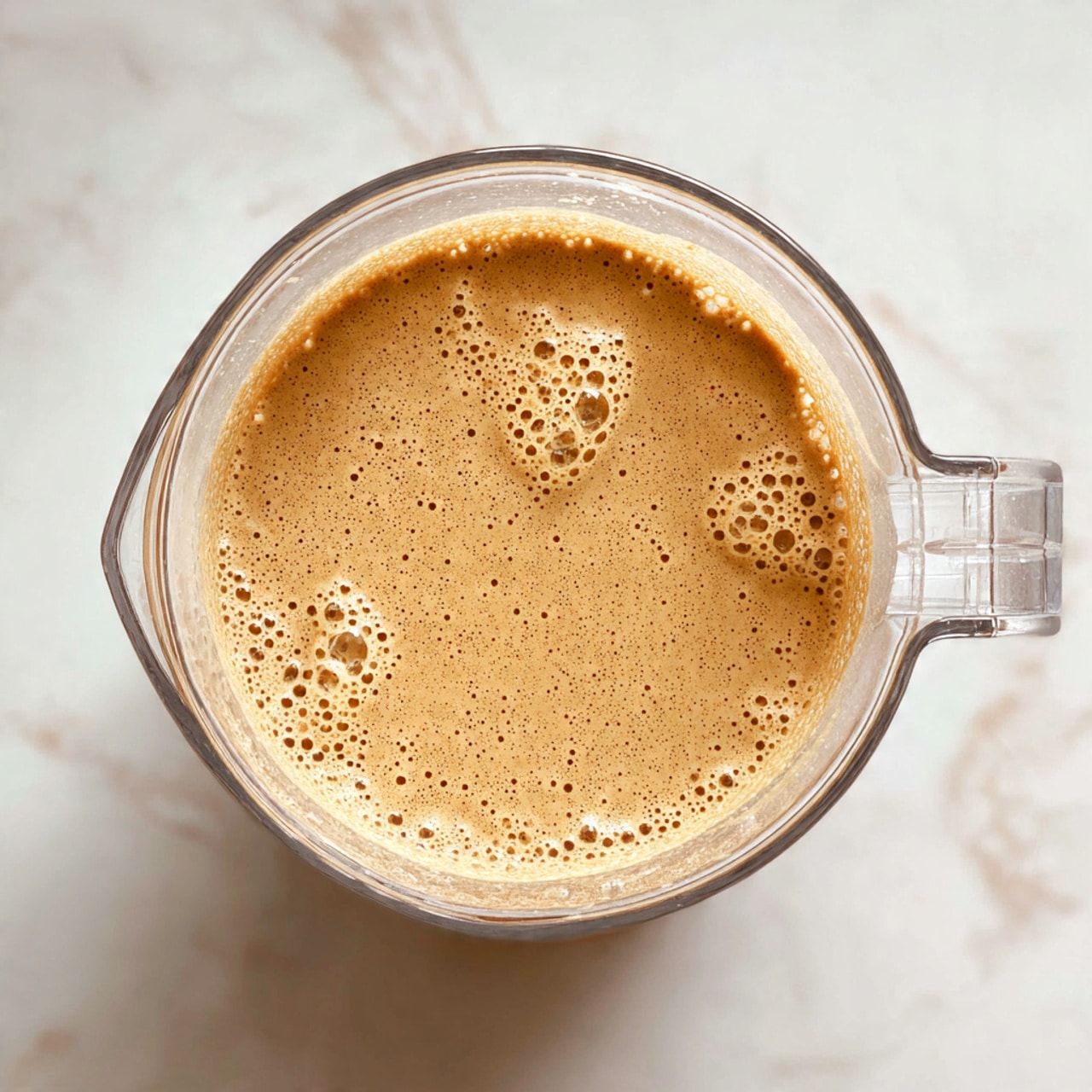 A top-down view of a clear blender container filled halfway with a smooth, light brown mixture that has small bubbles on its surface. The blender is placed on a white marbled textured surface, and the container's handle is visible on the right side. The mixture has a creamy texture with tiny darker specks spread evenly throughout photo taken with an iphone --ar 4:5 --v 7