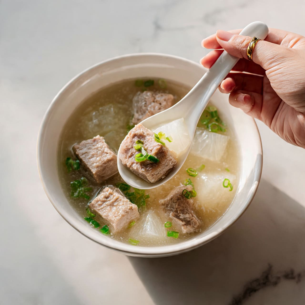 The image shows a white bowl filled with a clear soup placed on a white marbled surface. Inside the soup, there are three large pieces of light brown meat, along with translucent, pale white cubes that look like radish or turnip. Small green onion slices float on top, adding a bright green color contrast to the muted soup ingredients. A woman's hand holds a plain spoon, lifting some of the soup from the bowl. The overall look is clean and simple, with soft lighting that highlights the textures and colors in the bowl. photo taken with an iphone --ar 4:5 --v 7