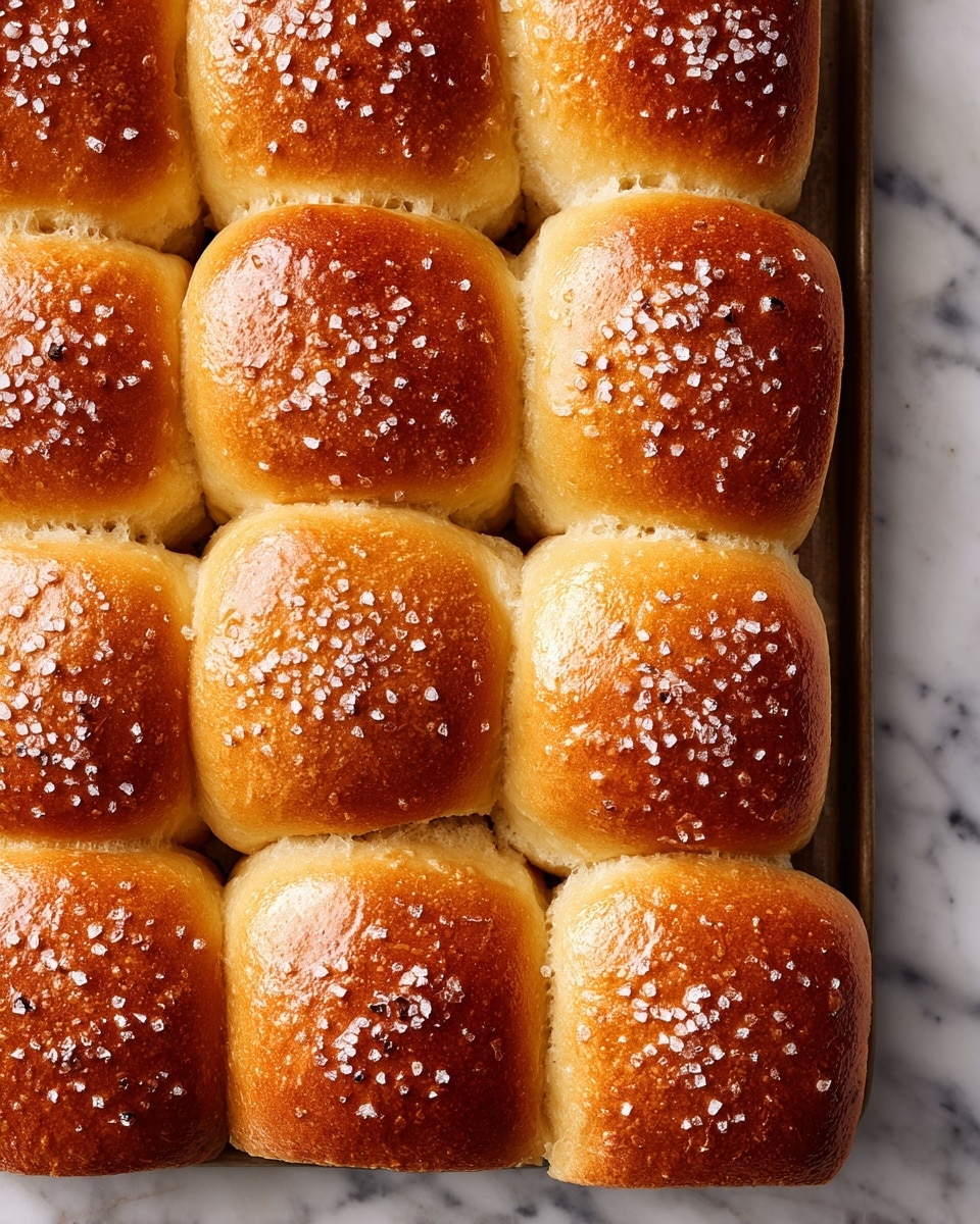 A close-up view of a tray filled with eighteen golden-brown bread rolls arranged in three rows of six. Each roll is perfectly puffed with a shiny, slightly textured surface topped with coarse salt crystals, giving them a warm, soft, and freshly baked look. The rolls are touching each other, creating a grid pattern with soft edges and a light crust. The background is a white marbled surface. Photo taken with an iphone --ar 4:5 --v 7