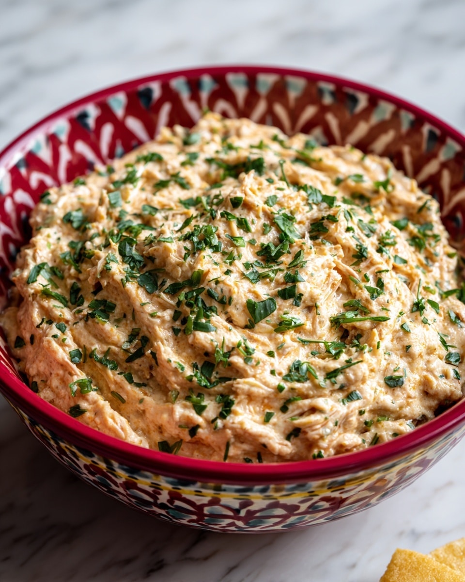 A red bowl with white, black, and yellow patterns holds a thick, creamy beige dish with visible bits of shredded meat or beans, topped with small pieces of chopped green herbs scattered evenly over the surface. The bowl is placed on a white marbled textured surface, with a small corner of a yellow chip visible at the bottom right. The creamy dish fills the bowl to the edges, showing a slightly uneven texture and a smooth yet chunky appearance. photo taken with an iphone --ar 4:5 --v 7