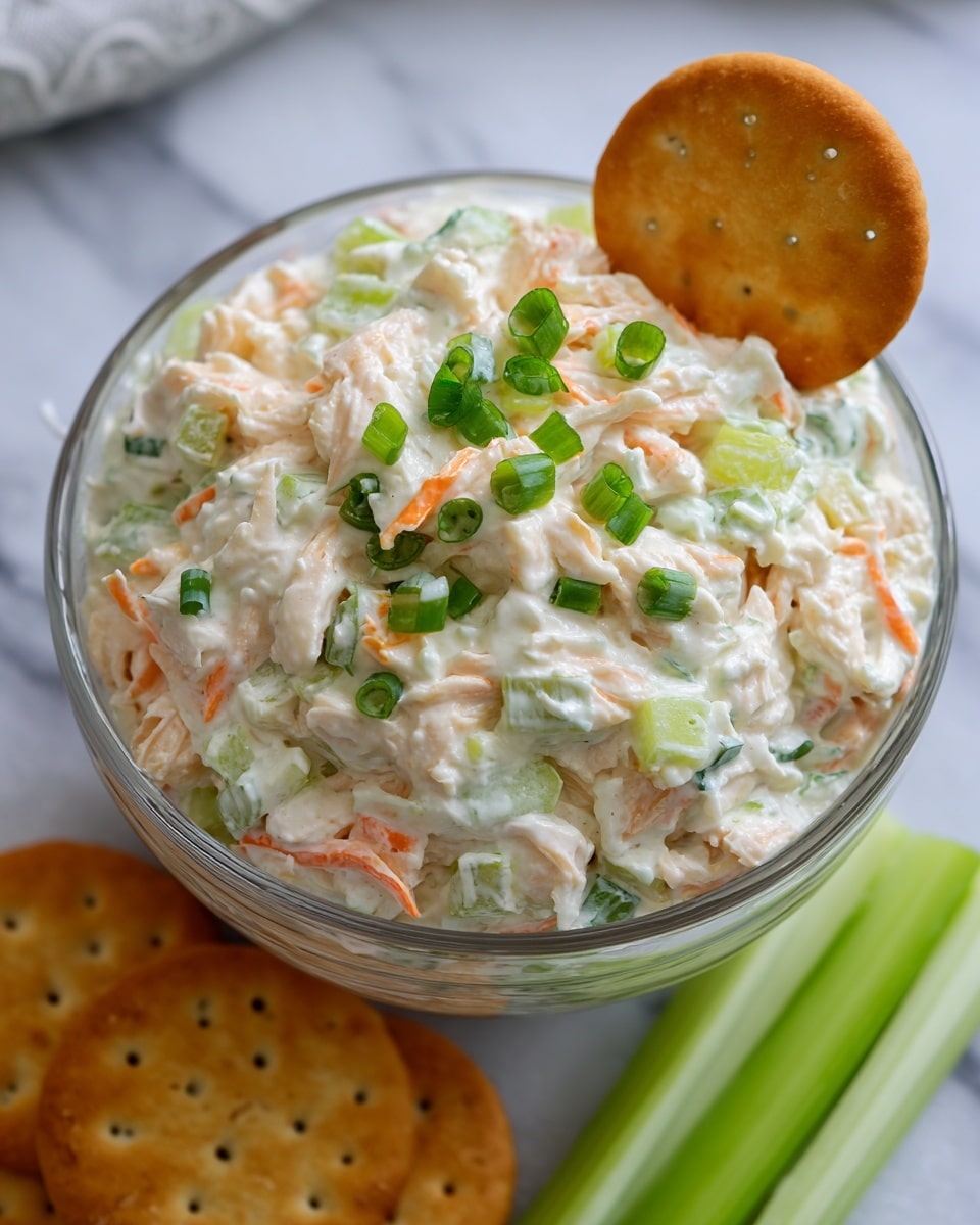 The image shows a clear glass bowl filled with a creamy white salad made of small chunks of chicken, orange carrot pieces, and pale green celery mixed smoothly together. The salad is topped with bright green chopped scallions sprinkled evenly on the surface. A round golden cracker is placed leaning on the side inside the bowl. Around the bowl on a white marbled surface, there are some more golden round crackers and two green celery sticks placed side by side. The overall look is fresh and inviting with soft creamy textures and pops of green and orange colors. photo taken with an iphone --ar 4:5 --v 7