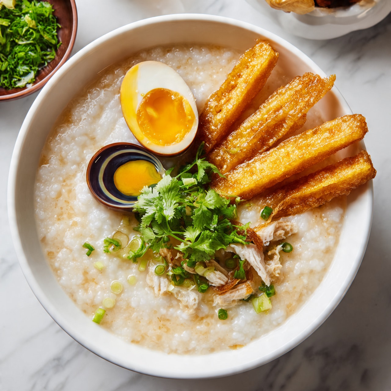 A white bowl filled with smooth, creamy rice porridge that has a light beige color. On top, there are several pieces of golden, crispy fried dough sticks positioned at the back right of the bowl. A shiny, dark century egg half with a translucent swirl of green and black is placed on the front left side. Scattered in the middle and front right are small pieces of cooked pale meat and fresh, bright green chopped scallions and cilantro leaves. The bowl sits on a white marbled surface, with a small bowl of green herbs blurred in the background. Photo taken with an iphone --ar 4:5 --v 7