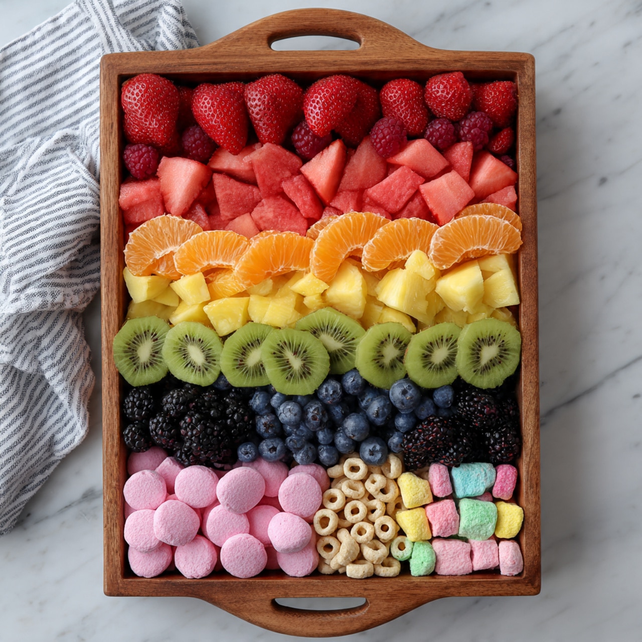 A wooden tray with a handle holds eight neat layers of food arranged from top to bottom: bright red halved strawberries with a smooth texture, small deep red raspberries, pink watermelon cubes, shiny orange mandarin segments, yellow pineapple chunks, bright green sliced kiwi, dark blue blueberries, and black blackberries with a bumpy surface. Below the fruit layers are two more rows: one of round pale pink candy pieces and the last one of tan cereal loops mixed with colorful marshmallow bits. The tray sits on a white marbled surface with a striped cloth partially visible on the left side. photo taken with an iphone --ar 4:5 --v 7