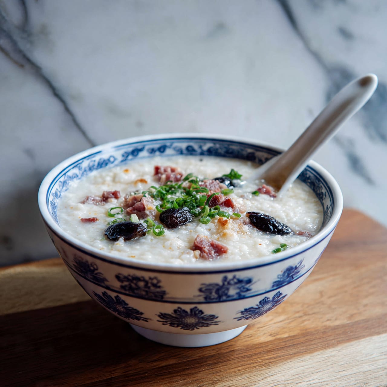 A white bowl holds a creamy, pale rice porridge base with a smooth texture, topped with several layers of garnishes. On one side, there are four golden-brown, crispy fried dough pieces, square and puffed. On the opposite side, a glossy, dark century egg half with a dark greenish-gray swirl sits partially submerged. Scattered on top are light brown pieces of tender meat and bright green chopped scallions and fresh herbs. The bowl is placed on a white marbled surface with a glimpse of a green vegetable bowl in the background. Photo taken with an iphone --ar 4:5 --v 7