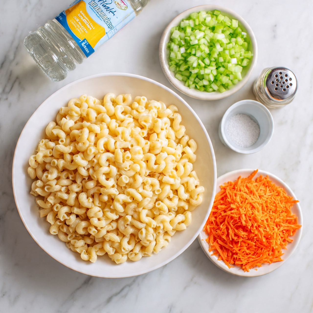 The image shows two white round plates and a small white bowl on a white marbled surface. The larger plate contains cooked elbow macaroni, light yellow in color with a soft texture, filling the plate. The smaller plate is divided into two piles: one of finely chopped green celery pieces on the top left, and one of grated bright orange carrot at the bottom center. Next to the celery, the small white bowl contains a clear liquid. A salt shaker and a bottle with a blue cap and yellow label are placed beside the plates. photo taken with an iphone --ar 4:5 --v 7