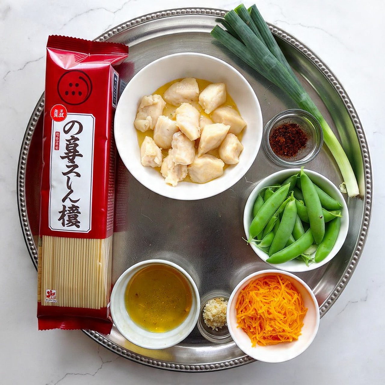 A silver tray holds various cooking ingredients neatly arranged. In the center is a white bowl filled with small pieces of light brown marinated chicken. To the right of it are two green onions resting diagonally, fresh and firm. Beside them is a small clear jar with dark brown and red spices inside. Below the chicken bowl, there is a white bowl full of fresh green snap peas, round and crisp. Next to the snap peas, on the bottom right corner, is a small clear bowl filled with bright orange shredded carrots. On the bottom left of the tray, there are two small clear bowls, one with a yellow liquid oil and the smaller one containing a minced light brown ingredient, likely garlic. On the left side of the tray lies a tall red package labeled