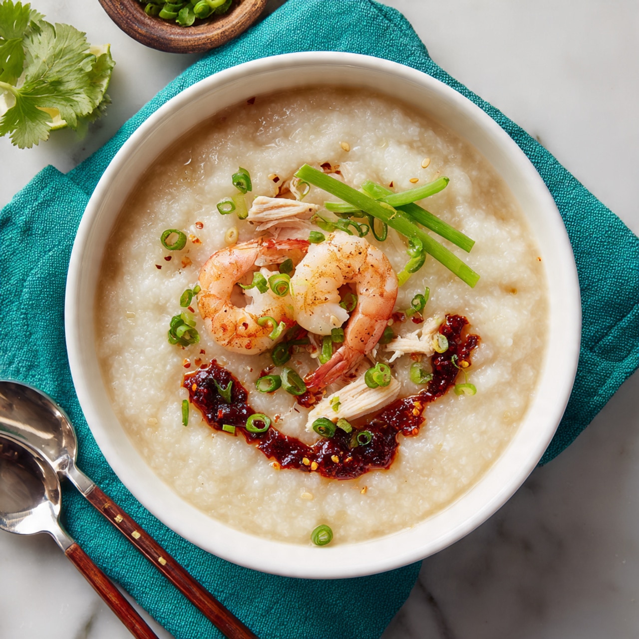 A white bowl filled with a creamy, light beige rice porridge base, topped with two pink and white shrimp placed near the center. There are a few pieces of shredded light brown chicken beside the shrimp. Thin bright green strips of scallion rest on one side, while dark red chili oil with small bits of chili forms a circle around part of the porridge near the edge. Small green herb pieces are scattered lightly on top. The bowl sits on a white marbled surface with a turquoise cloth partially under it, with a lime wedge, cilantro leaves, a silver spoon, and wooden chopsticks nearby. Photo taken with an iphone --ar 4:5 --v 7