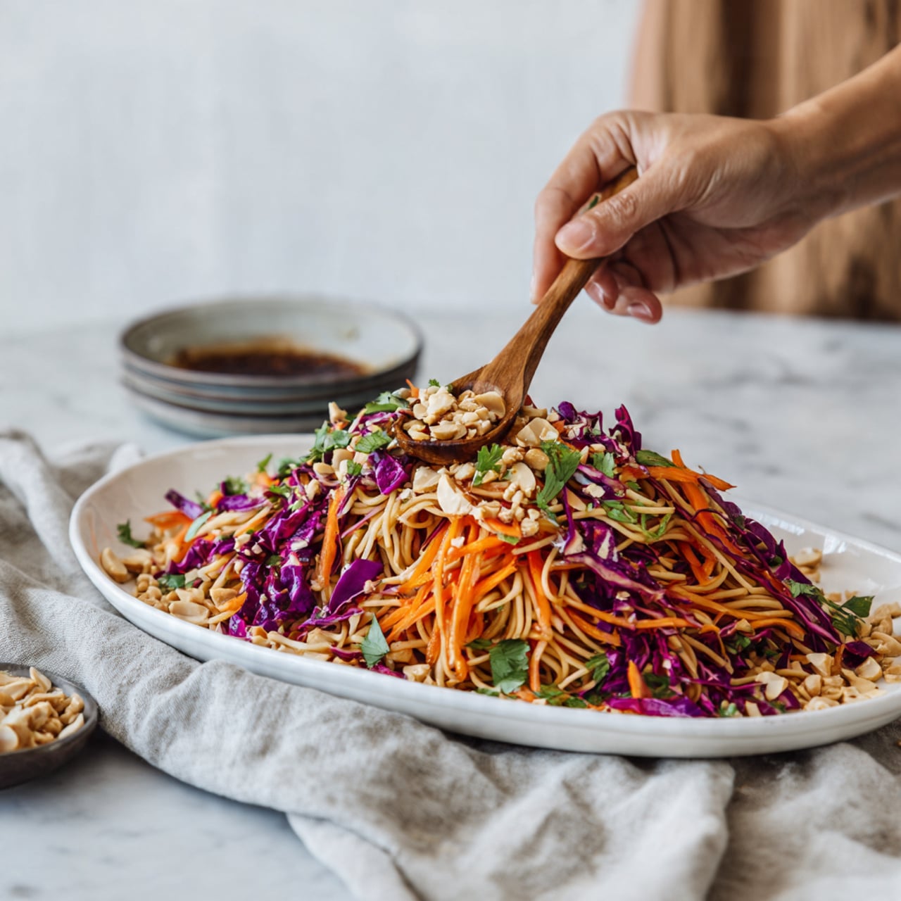 The image shows a long white plate filled with a noodle salad. The bottom layer is light brown noodles, twisted and spread evenly. On top of the noodles, there are colorful layers of thin orange carrot strips, purple cabbage, and green herbs scattered across. Light tan crushed peanuts are sprinkled over the entire dish. The plate is placed on a white marbled surface with a light gray cloth nearby and a small bowl of dark sauce visible in the top left corner. A woman's hand holding a wooden spoon is adding the noodles. photo taken with an iphone --ar 4:5 --v 7