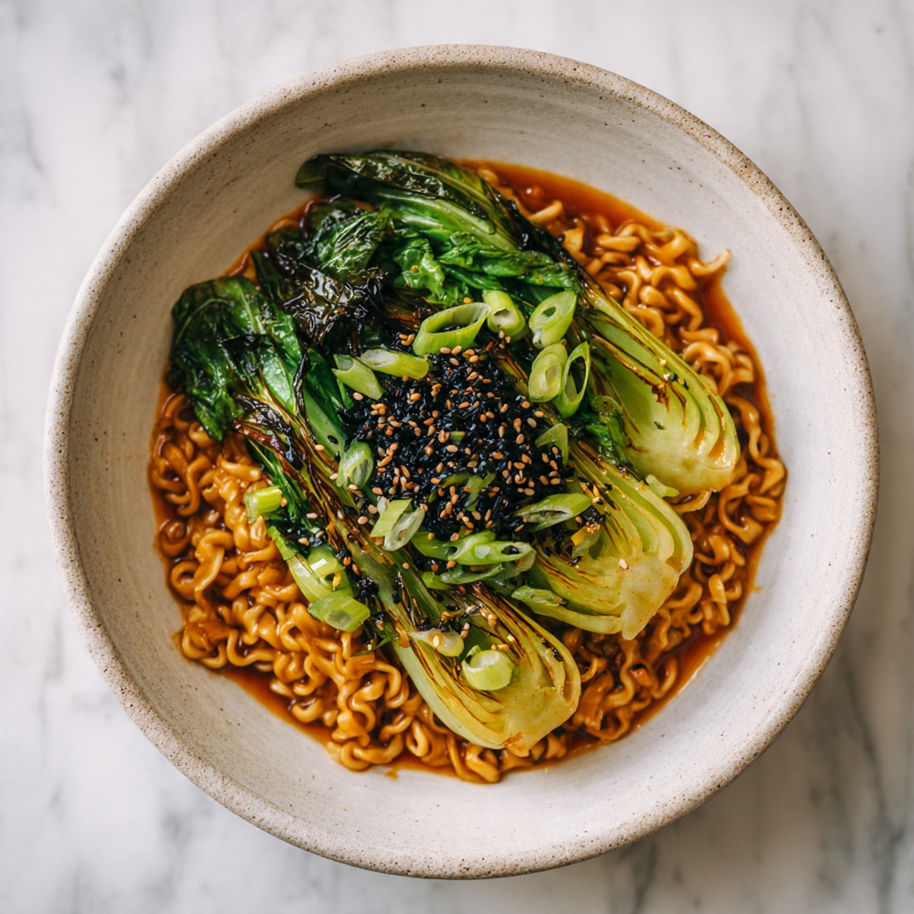 The image shows a bowl of thick, golden-brown noodles coated with a shiny sauce forming the bottom layer. On top of the noodles, there is a layer of bright green cooked bok choy with a slight char, adding texture and a fresh look. The dish is garnished with thinly sliced spring onions scattered around the bok choy, and a generous sprinkle of black and white sesame seeds sits in the center, giving contrast and detail. The bowl itself is white with a natural stone texture, placed on a white marbled surface. photo taken with an iphone --ar 4:5 --v 7