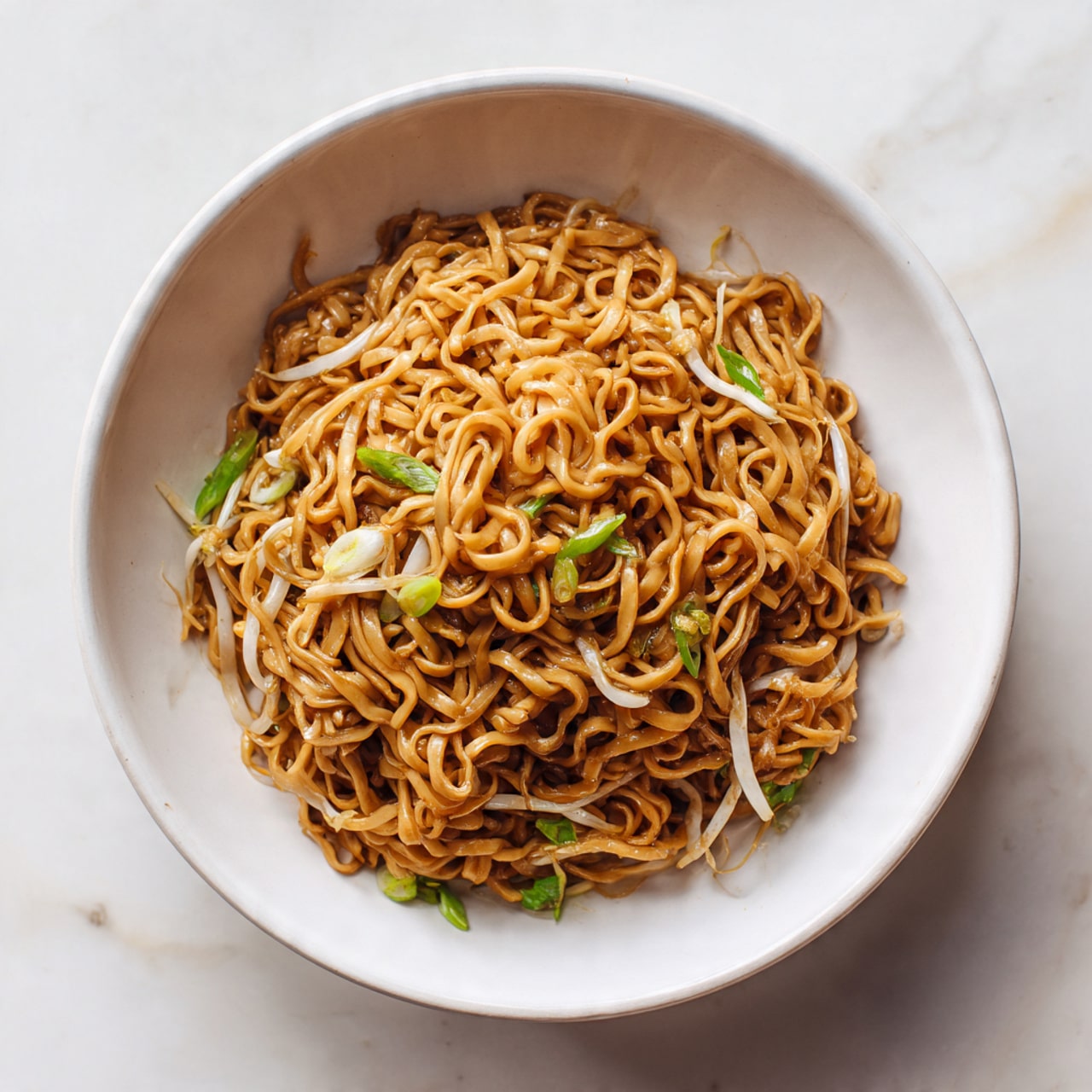 A white bowl filled with a single thick layer of brown noodles twisted into a small pile, mixed with thin white bean sprouts and small green onion pieces spread evenly throughout. The noodles have a slightly shiny, smooth texture, coated with a sauce that gives them a warm brown color. The bowl sits on a white marbled surface with soft lighting highlighting the colors and details of the noodles and vegetables photo taken with an iphone --ar 4:5 --v 7