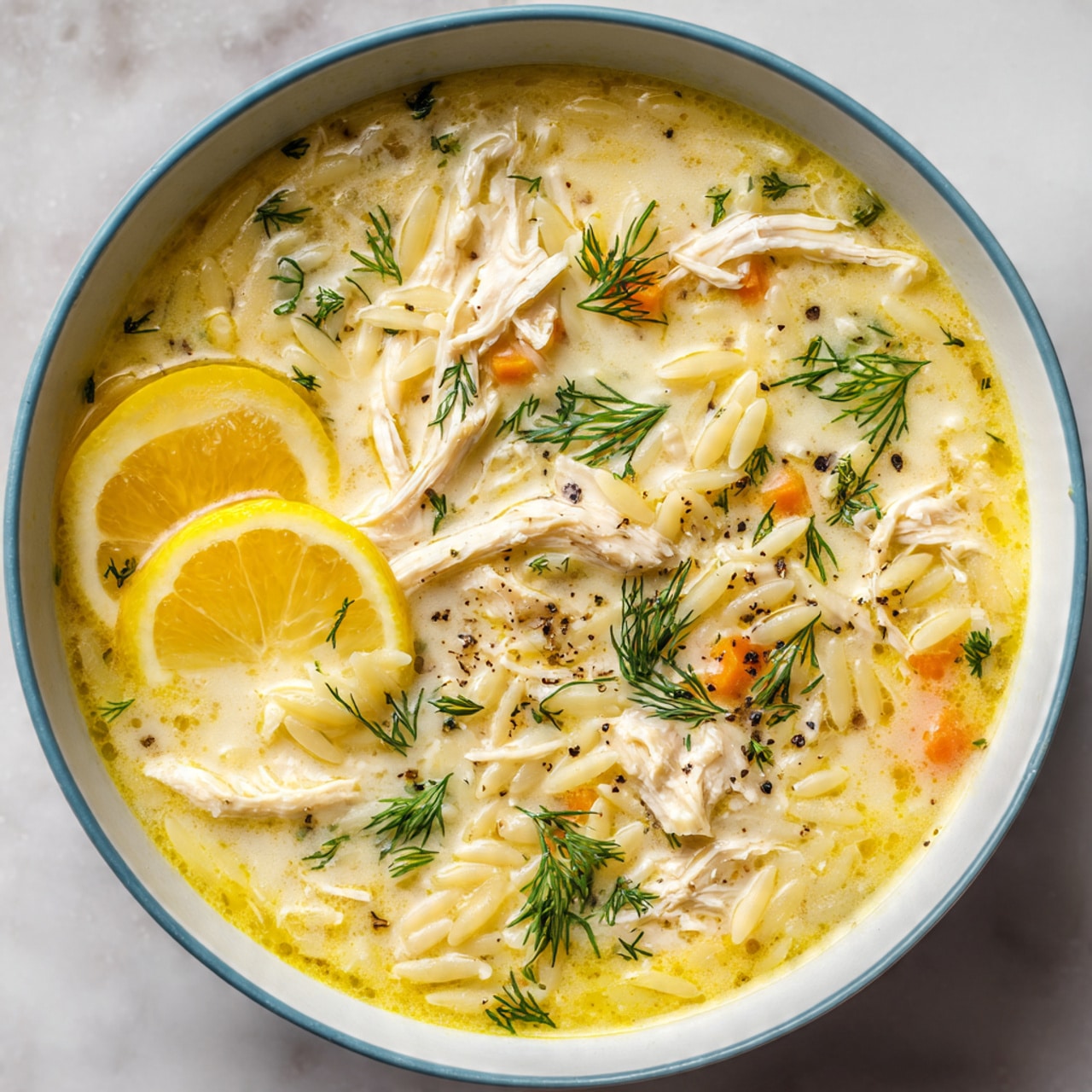 A close-up of a light yellow chicken soup in a white bowl with a light blue rim. The soup has shredded chicken pieces scattered throughout with some white orzo pasta mixed in. There are two thin lemon slices floating near the bottom left side of the bowl. Small sprigs of fresh green dill are spread across the surface, along with a few specks of black pepper. Tiny oil droplets can be seen on the soup’s surface, adding a slight shine. The bowl is placed on a white marbled textured surface photo taken with an iphone --ar 4:5 --v 7