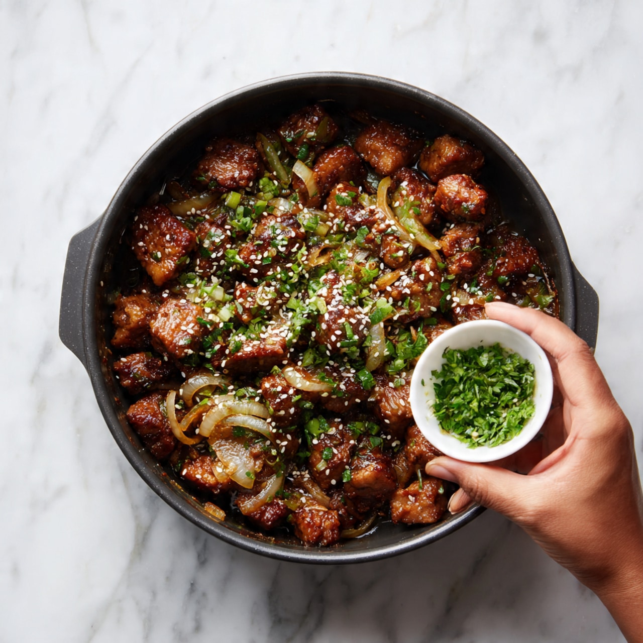 The image shows a close-up of a black pot filled with cooked brown cubed pieces that look tender and glossy, mixed with translucent cooked onions. The dish is topped with small white sesame seeds and chopped green herbs evenly spread across the surface. On the right side, a woman's hand is holding a small white bowl, pouring fresh green herbs into the pot. The pot rests on a white marbled surface. photo taken with an iphone --ar 4:5 --v 7