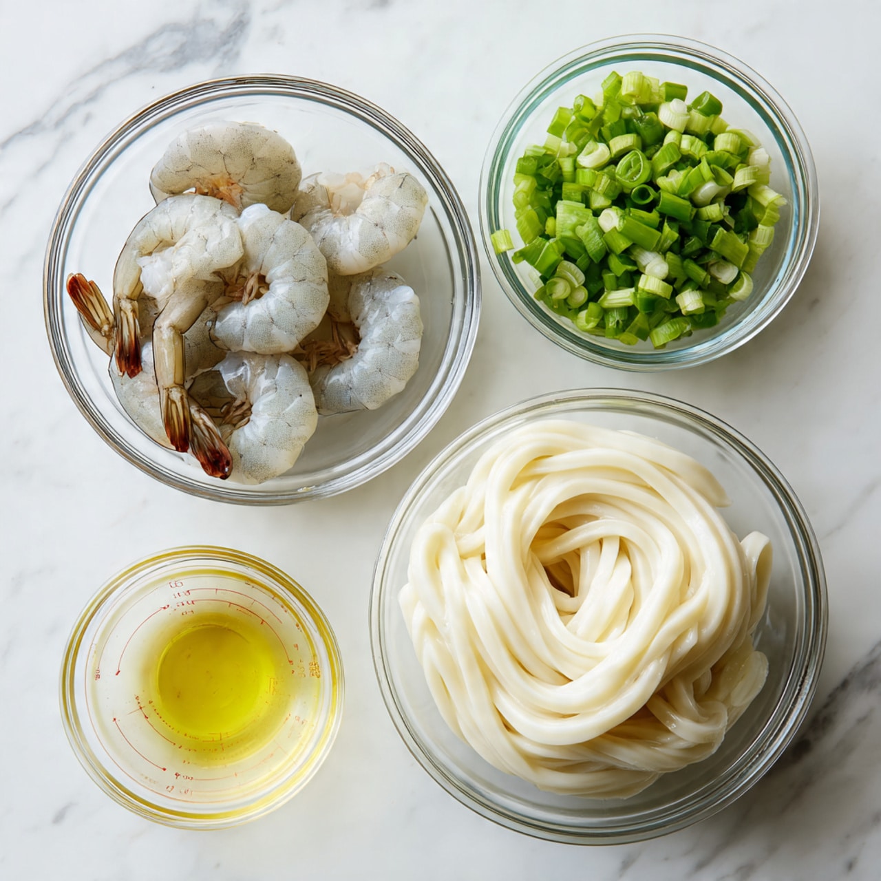 The image shows four clear glass bowls arranged on a white marbled surface. The largest bowl in the bottom right contains a block of thick, white udon noodles with a smooth texture, coiled loosely inside. To the upper left, a medium bowl holds five raw, peeled shrimp with a light grayish-white color and slightly translucent bodies, their brown tails still attached. Above the shrimp, a small bowl is filled with bright green chopped scallions, giving a fresh, crisp look with varied green shades. To the right of the scallions, a tiny bowl contains a pale yellow liquid, likely cooking oil, with a smooth, glossy surface and small measurement marks visible on the side of the bowl. Photo taken with an iphone --ar 4:5 --v 7