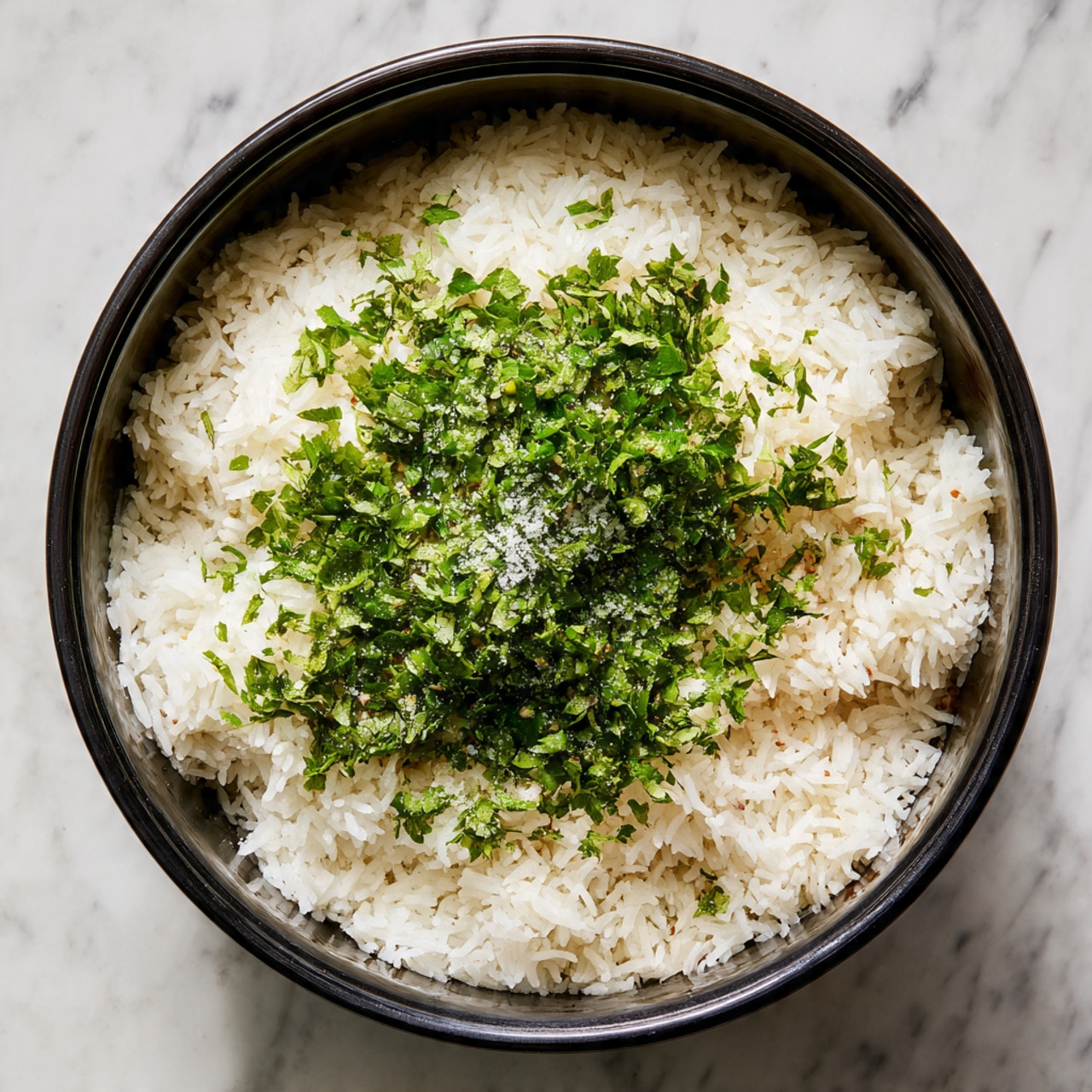 Inside a black pot, there is a thick base layer of fluffy white rice with a soft texture. On top of the rice, in the center, there is a thick layer of finely chopped fresh green herbs, scattered evenly but piled up slightly in the middle. The herbs have a rough texture and bright green color. The light shines from above, highlighting the grains of rice and the leafy herbs, with some coarse salt visible on the herbs. The pot sits on a surface with a white marbled texture. Photo taken with an iphone --ar 4:5 --v 7