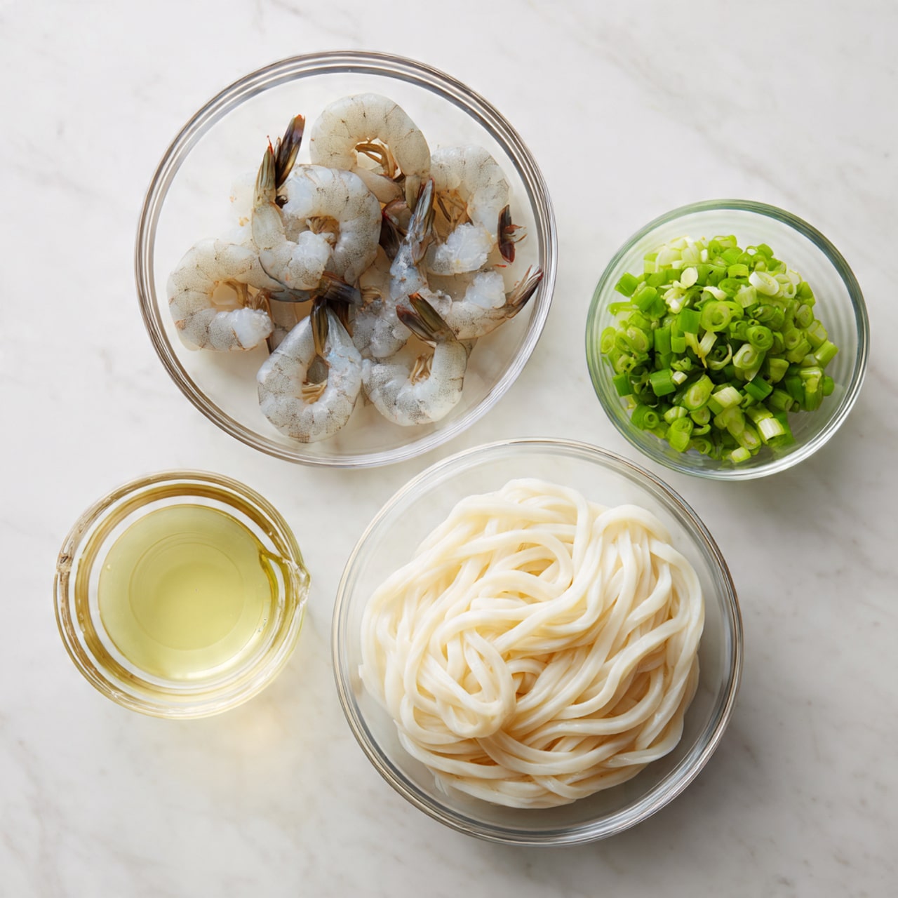The image shows four clear glass bowls arranged on a white marbled surface. The largest bowl at the bottom right contains a block of thick, white udon noodles with a smooth, slightly shiny texture. Above it to the left, another glass bowl holds five raw shrimp, pale gray with translucent shells and darker tails, curled and resting flat in the bowl. To the upper right of the shrimp bowl, a small glass bowl contains bright green chopped scallions, finely cut with a fresh, crisp look. To the far right, a small clear measuring cup holds a pale yellow liquid, likely oil or broth. The setup is simple and clean, with all items clearly visible and well-lit, photo taken with an iphone --ar 4:5 --v 7