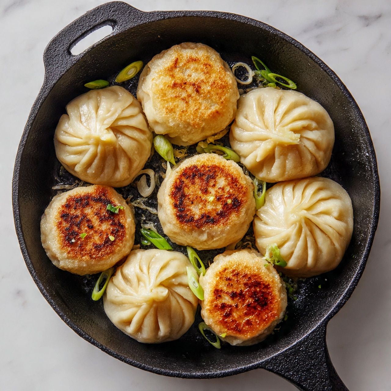 The image shows a black cast iron pan on a white marbled surface filled with eight round dumplings arranged in a circle. Seven dumplings are pale beige with pleated, pinched tops and a soft, doughy texture, while one dumpling has a golden brown crispy bottom facing up, showing a toasted crust. A few small green sliced scallions are scattered across the dumplings and pan, adding a touch of color. The pan's handle is visible at the top right. Photo taken with an iphone --ar 4:5 --v 7