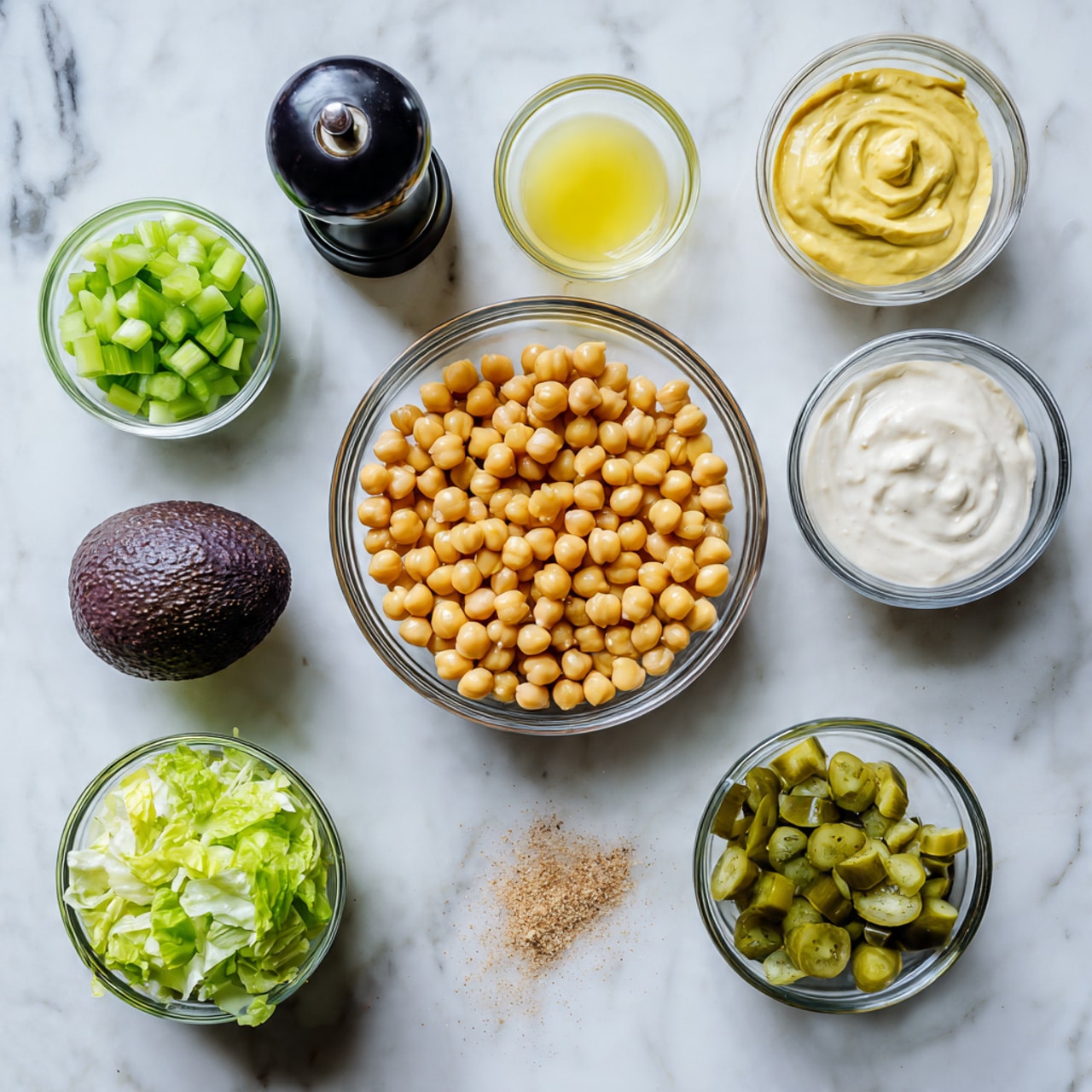 The image shows several glass bowls and a black pepper grinder arranged neatly on a white marbled surface. In the center, there is a medium-sized glass bowl filled with yellow chickpeas. Surrounding it are smaller glass bowls containing different ingredients: chopped green celery at the top left, a light yellow liquid (likely lemon juice) at the top center, a bright yellow spread (likely mustard) at the top right, creamy white yogurt or sauce at the middle right, and chopped green pickles at the bottom right. At the bottom left, a small glass bowl contains torn green lettuce leaves. Below the chickpeas, there is a small pile of a light brown spice on the marble surface, and near it sits a dark purple avocado. The black pepper grinder is positioned on the left side of the image. The photo is taken with a woman's hand not visible in this shot photo taken with an iphone --ar 4:5 --v 7