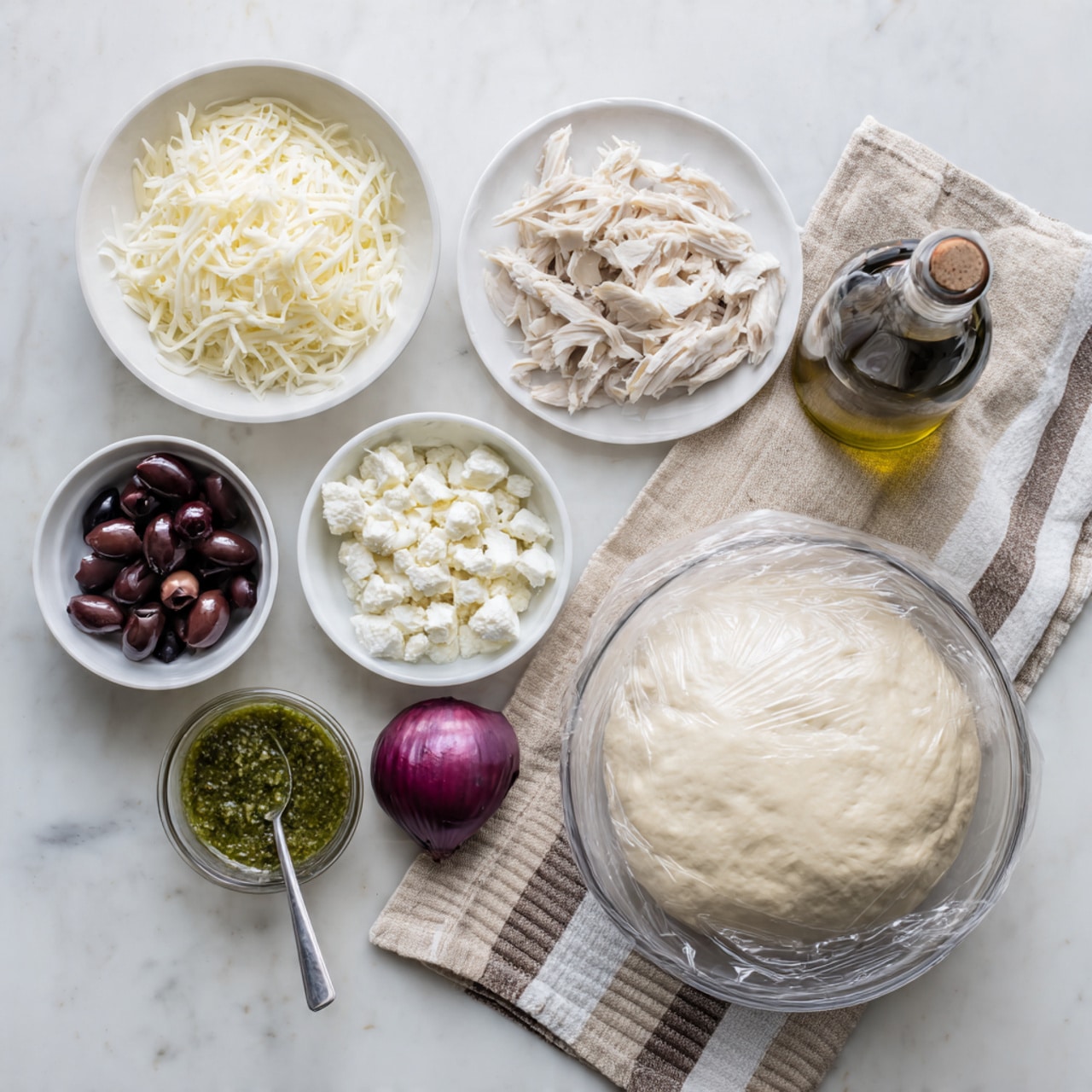A flat lay image shows ingredients ready for cooking on a white marbled surface. There is a large clear glass bowl on the right filled with soft, risen dough covered with plastic wrap. Below it, a small white bowl holds finely chopped dark purple olives. To the left, a white plate contains shredded white chicken meat. Above the chicken, a white bowl is filled with shredded white cheese. Next to it, a white bowl has whole dark olives. Another small white bowl contains crumbly white cheese. A halved red onion sits beside the bowls. In the center is a small clear plastic container holding green pesto sauce, with a silver spoon resting inside. A dark bottle of extra virgin olive oil is placed upright on the far right. All items are arranged neatly on a folded beige and white striped cloth. Photo taken with an iphone --ar 4:5 --v 7