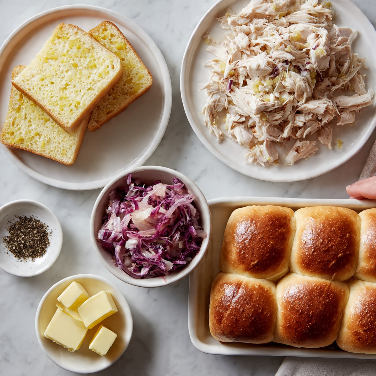 The image shows four white dishes on a white marbled surface. In the top left is a white plate with three slices of yellowish bread stacked neatly. To the right, on a white plate, is a pile of shredded light brown and white chicken. Below the bread, there is a white bowl filled with a chunky purple sauce or salad. Next to it, a small white dish holds a pile of black pepper. At the bottom left, a small white dish contains several slices of yellow butter. On the right side, a larger white dish holds a grid of square golden brown bread rolls with smooth, slightly shiny tops. A woman's hand is reaching in from the right side, touching the bread rolls. The photo taken with an iphone --ar 4:5 --v 7